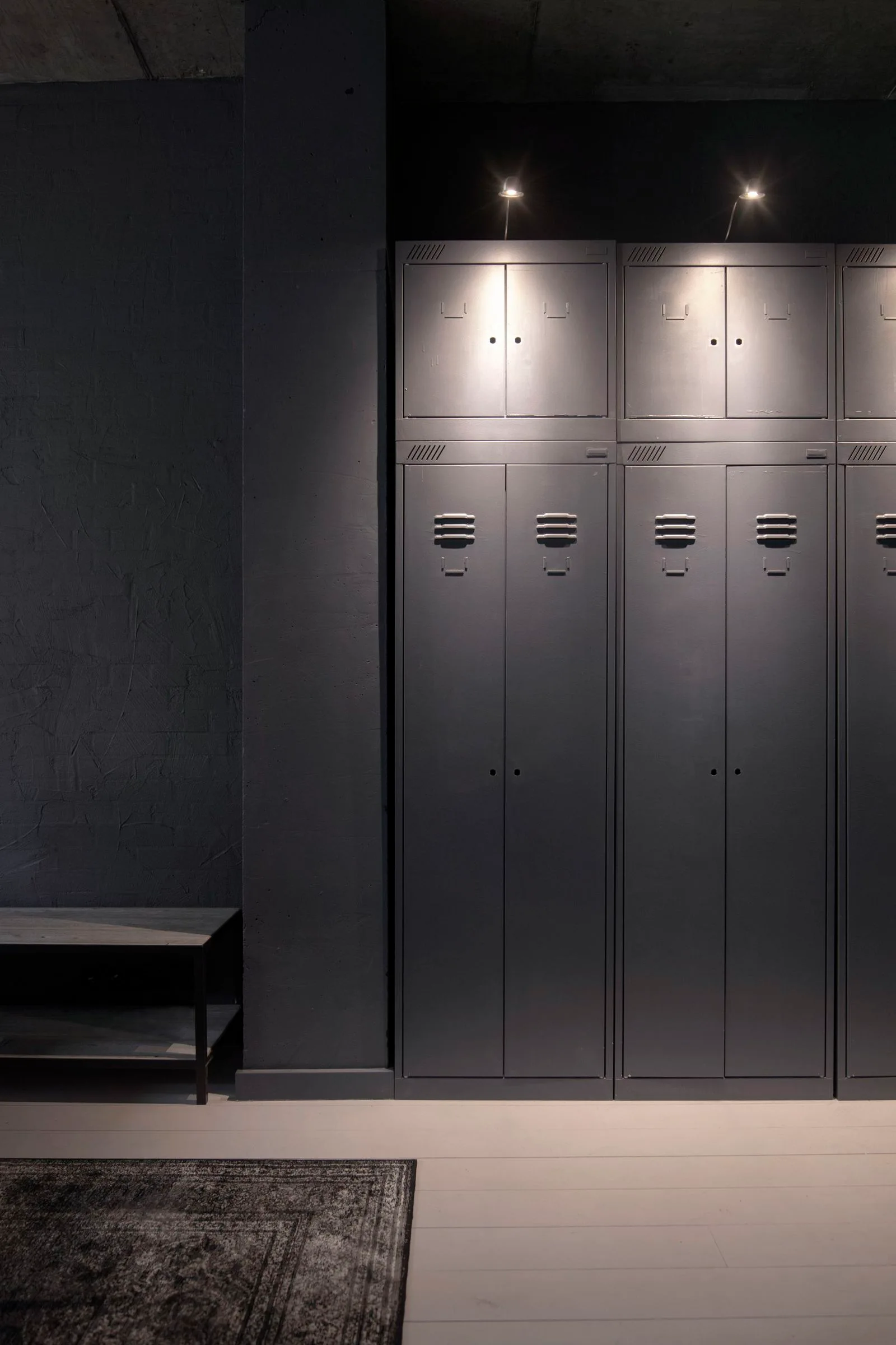 Tall grey metal storage lockers with vents stand next to a charcoal concrete pillar and a low wooden bench under soft spotlights.
