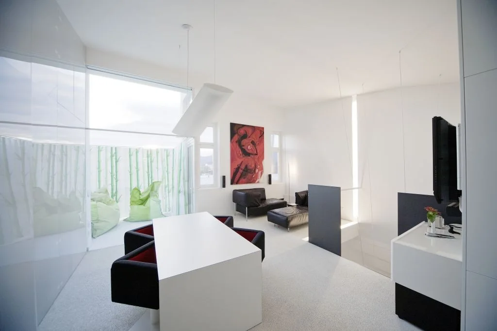 A minimalist open-plan apartment with white walls, a white dining table, black seating, and a large red abstract painting on the wall.