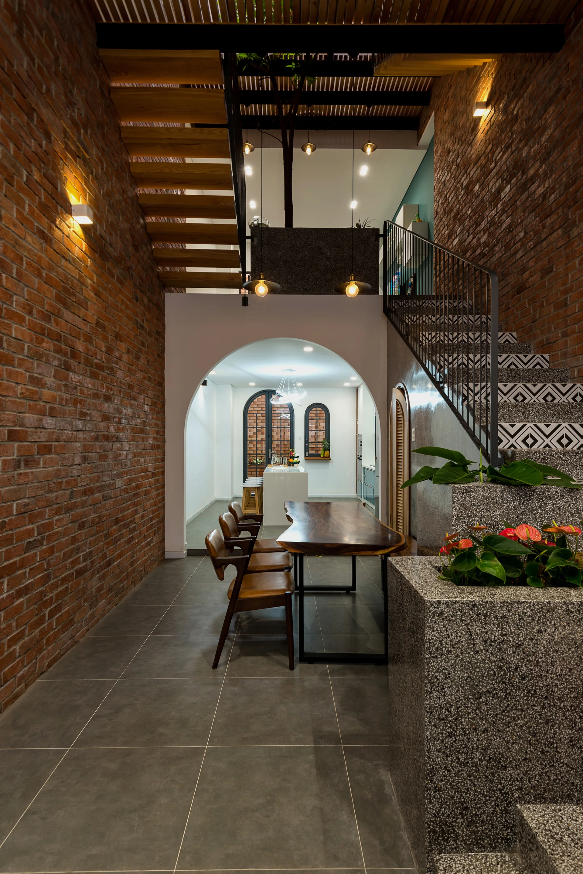 A modern dining room features a live-edge wood table beneath exposed brick walls, with a tiled staircase and an arched opening to a kitchen.