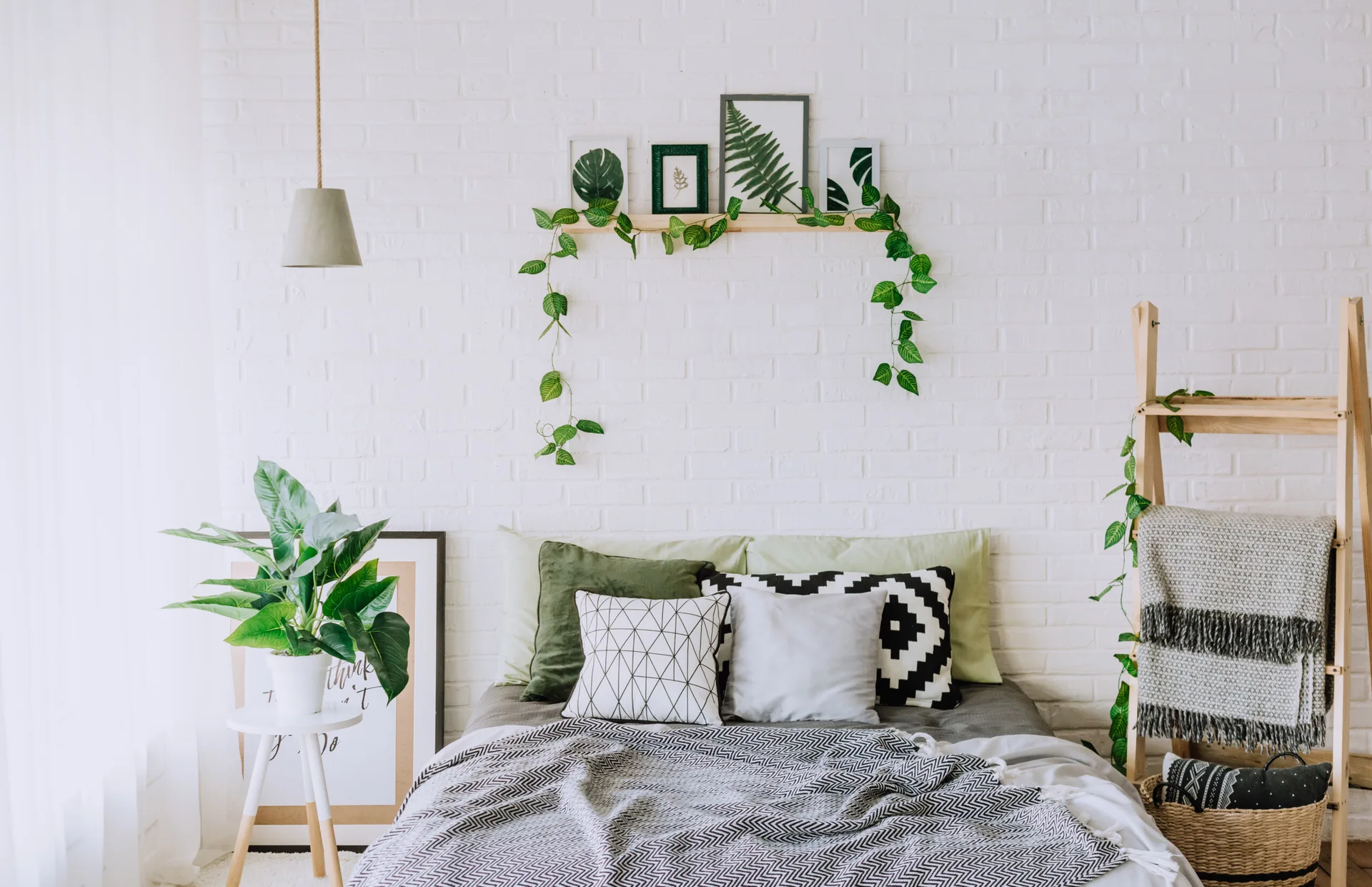 A bedroom features a bed with green and geometric pillows set against a white brick wall, decorated with a floating shelf, plants, and a wooden ladder.