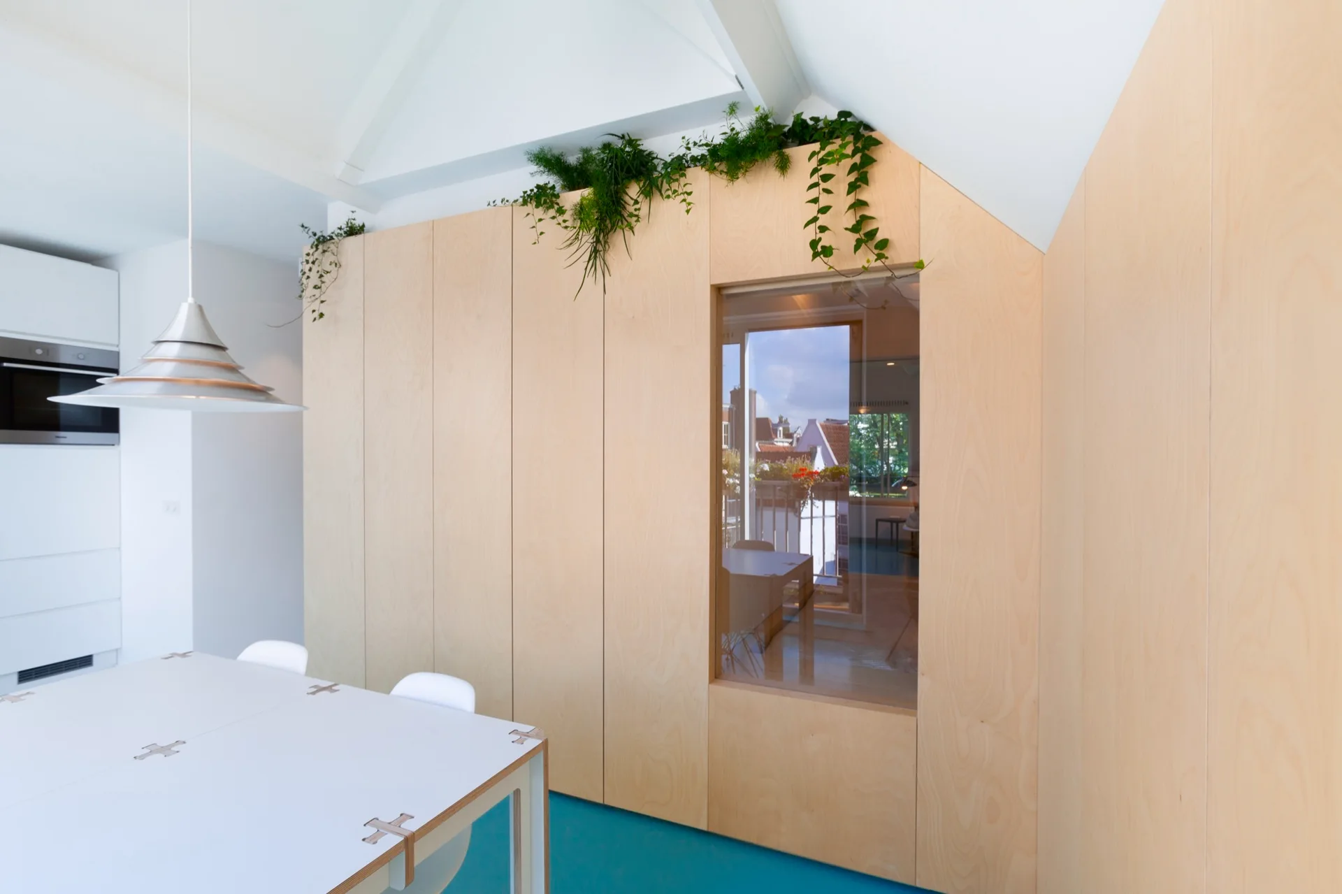 A modern dining room featuring a floor-to-ceiling plywood cabinet wall with a built-in window, a white dining table, and blue flooring.