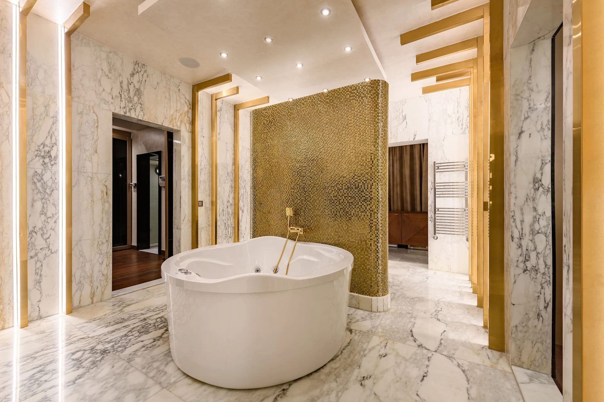 A white freestanding bathtub sits in front of a curved gold mosaic partition wall in a bathroom lined with veined white marble.