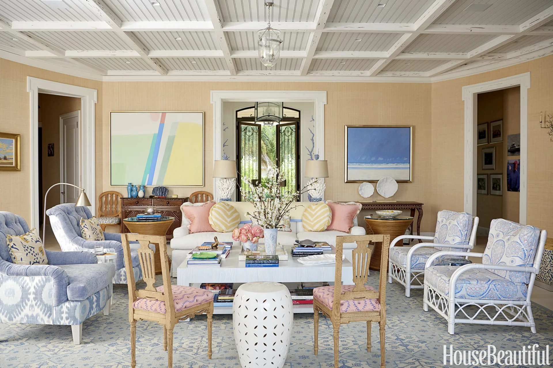 A coastal living room features a white sofa, patterned blue armchairs, a white coffee table, grasscloth walls, and a white paneled coffered ceiling.