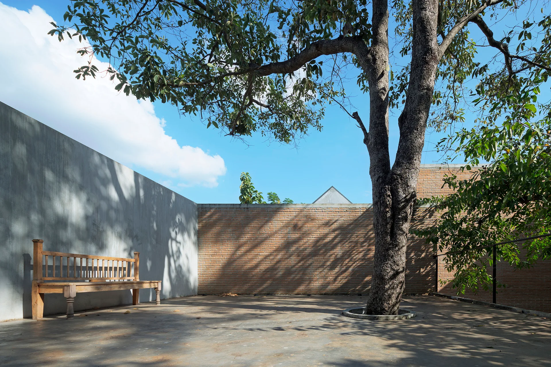 A spacious outdoor courtyard featuring a wooden bench against a concrete wall, surrounded by brick walls under a blue sky.
