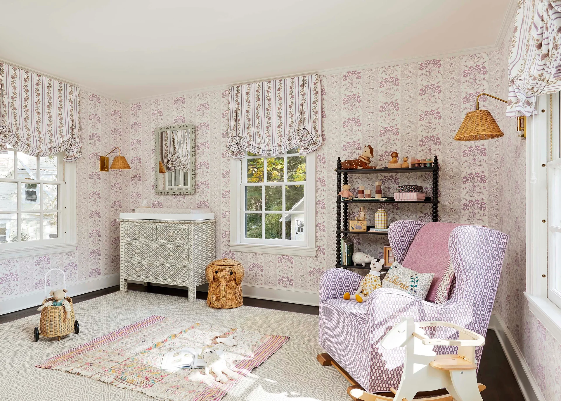 A nursery with purple floral wallpaper, a patterned wingback rocking chair, a white bone inlay dresser, and a black spindle bookshelf.