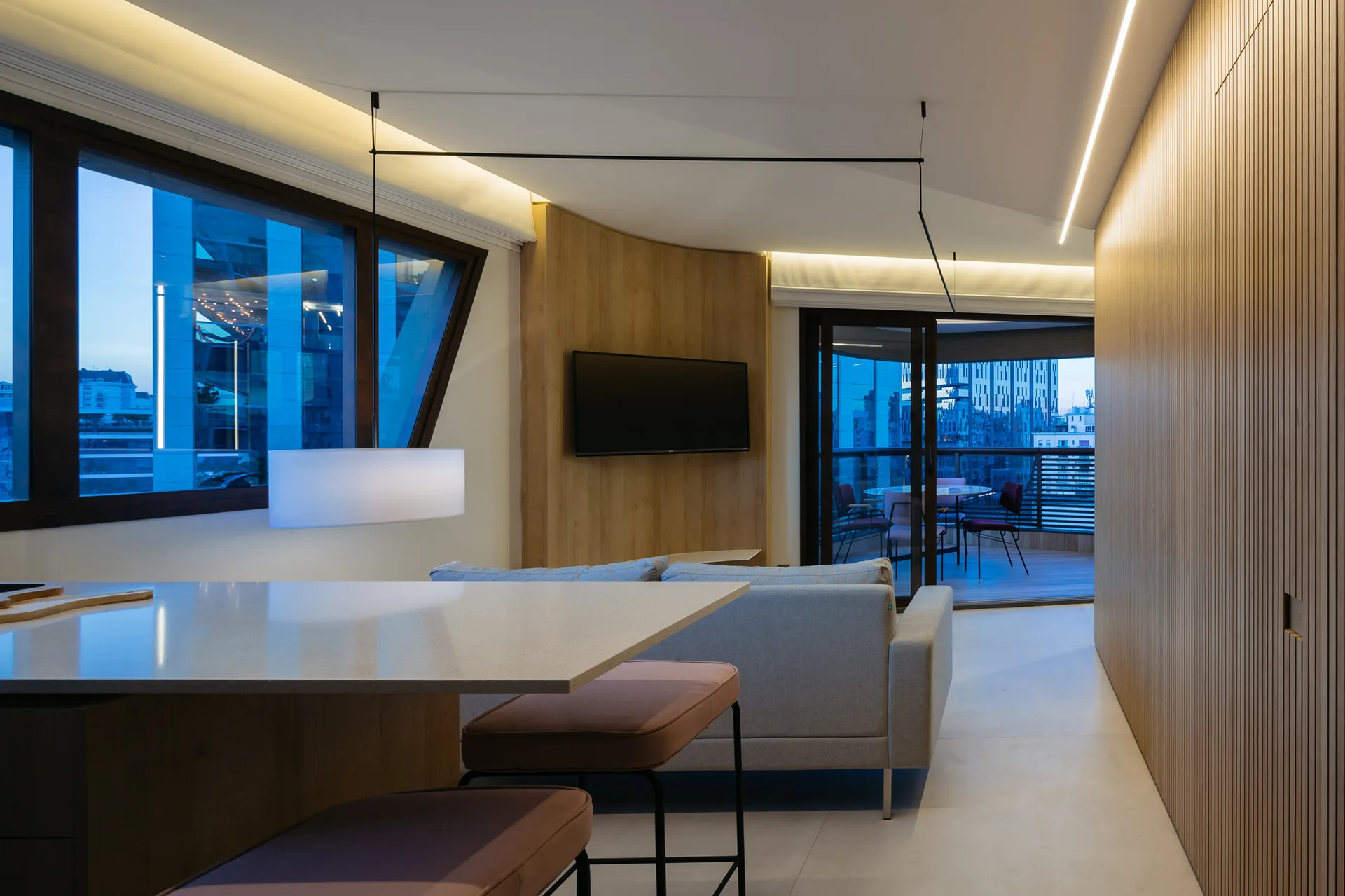 A modern open-plan living room features a grey sofa, kitchen island with stools, and vertical slatted wood walls beside large windows at dusk.