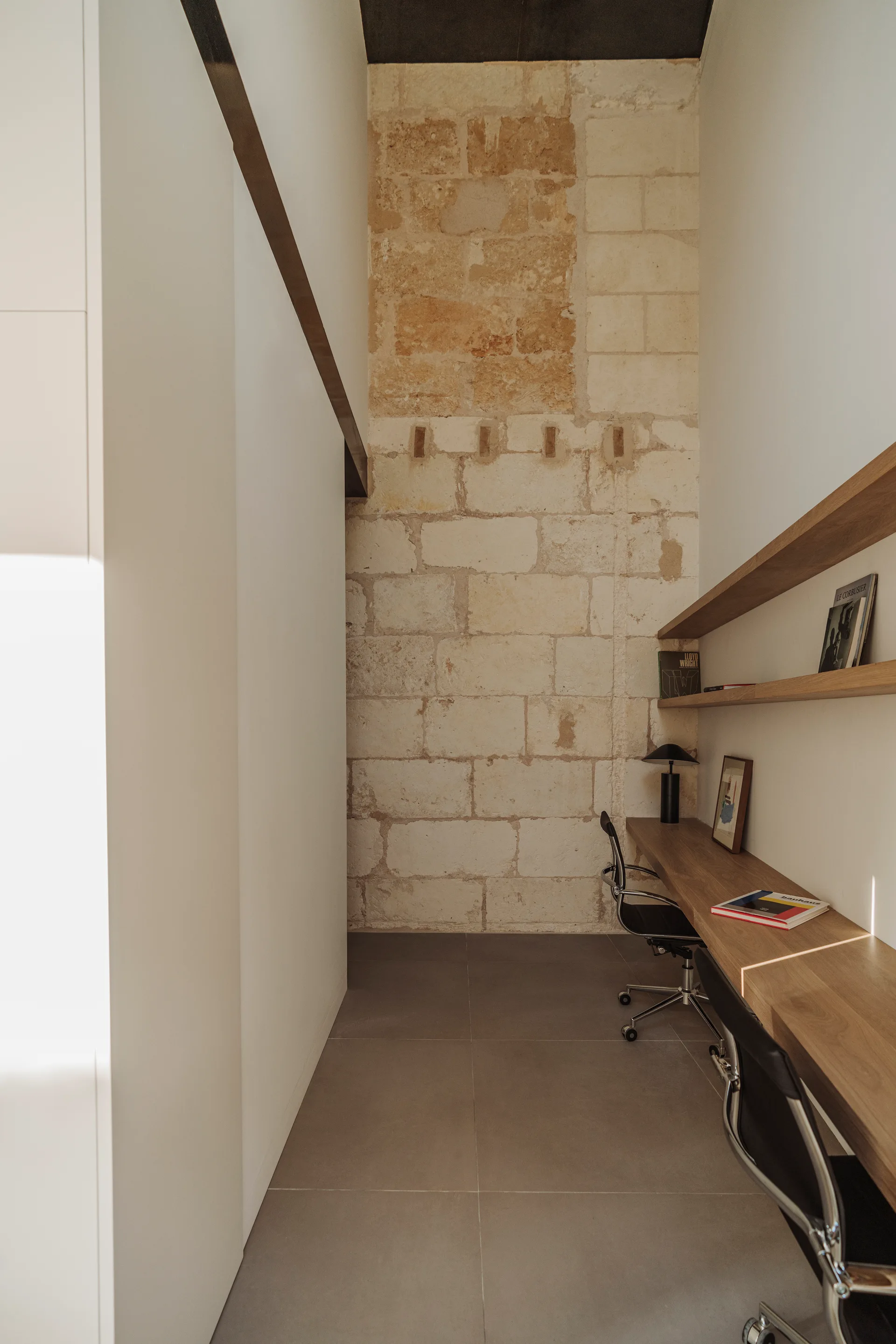 A narrow home office features a long wooden desk, floating shelves, black office chairs, and an exposed stone wall under bright natural light.