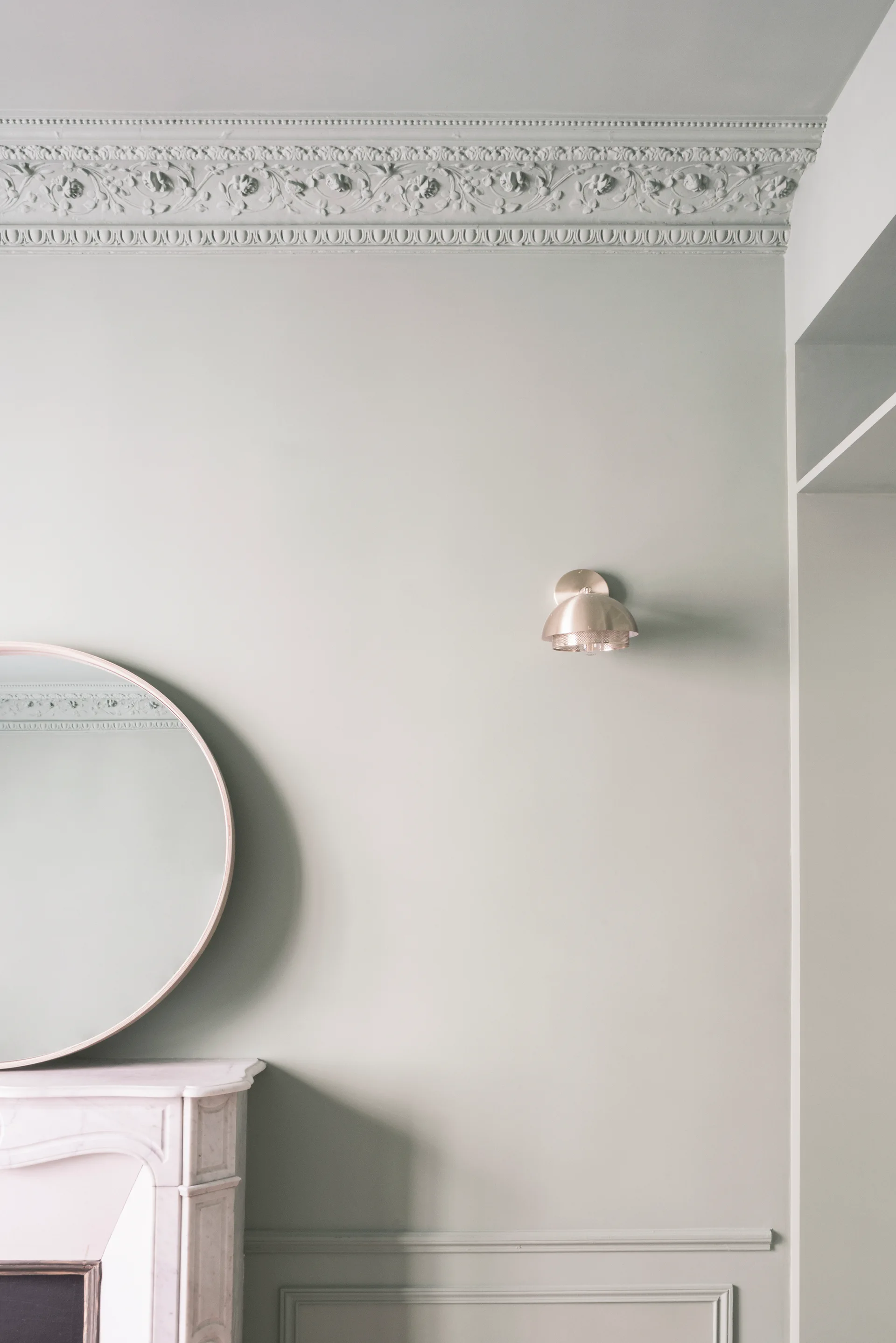 A pale sage green wall featuring ornate floral plaster crown molding, a brass dome sconce, and a round mirror on a white marble fireplace mantel.