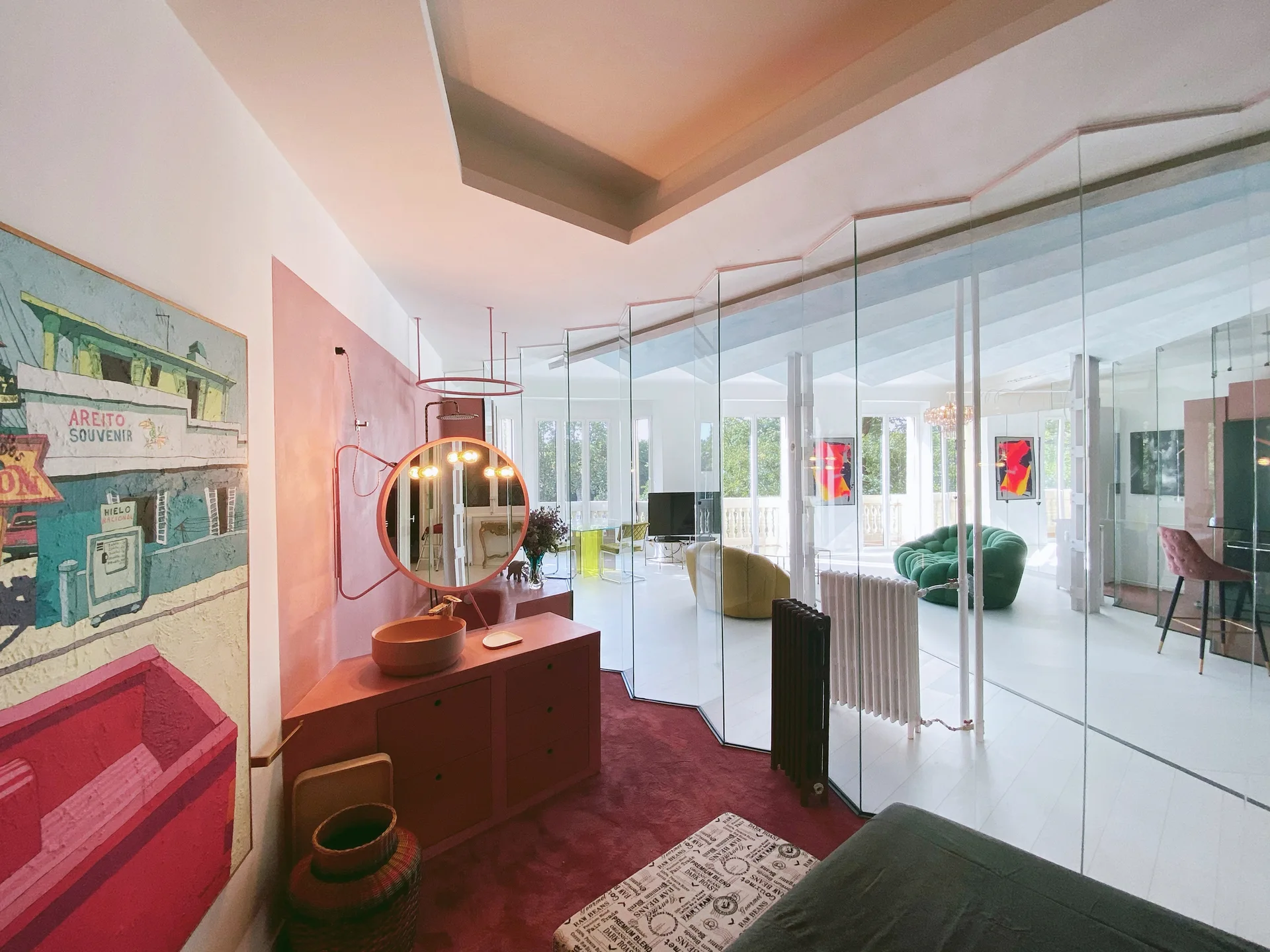 A pink dressing area with a built-in vanity and round illuminated mirror separated from a bright living space by angled glass partitions.