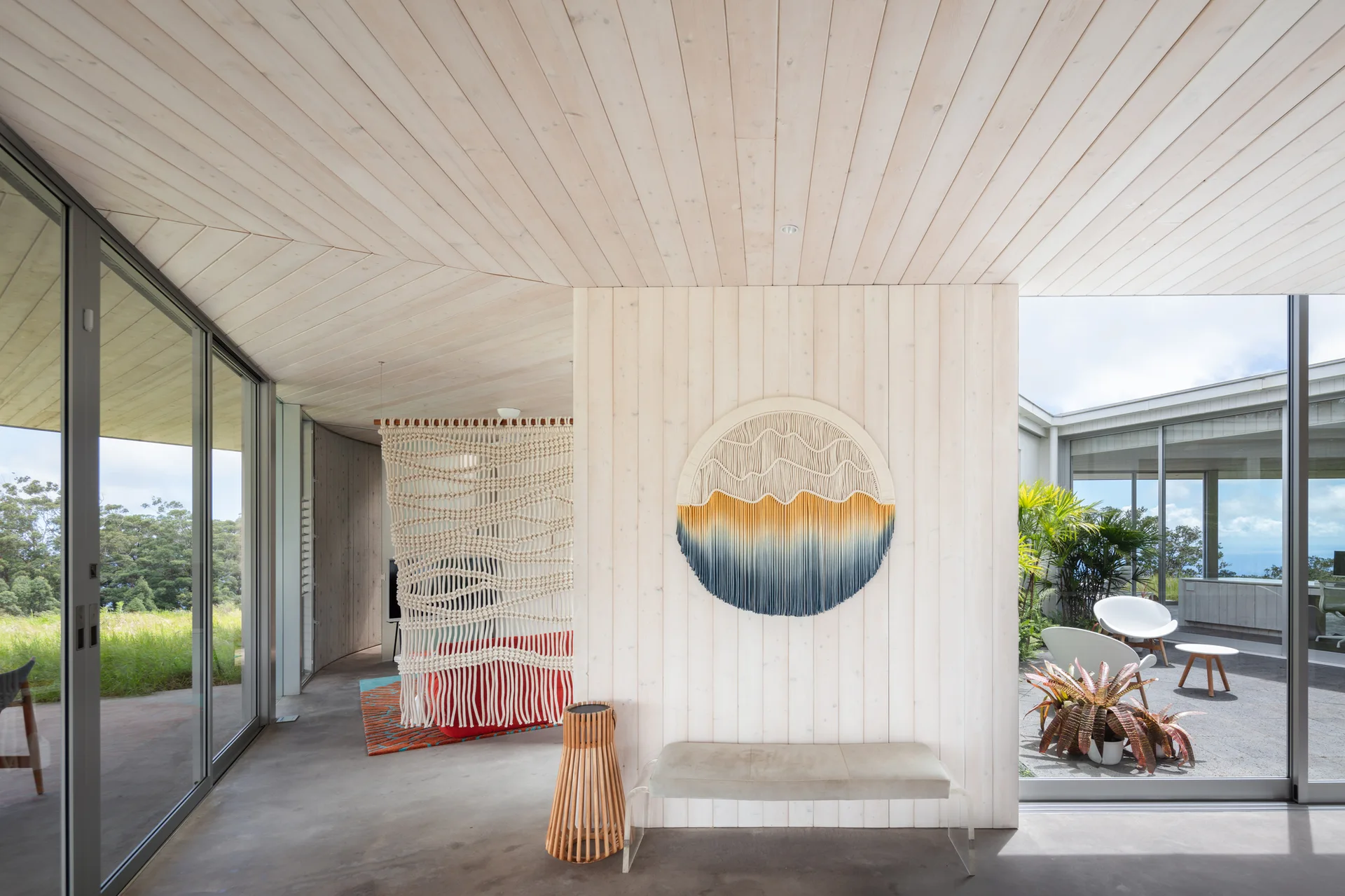 A modern interior space features light wood-paneled walls and ceiling, a circular woven wall hanging, and a low bench with clear acrylic legs.