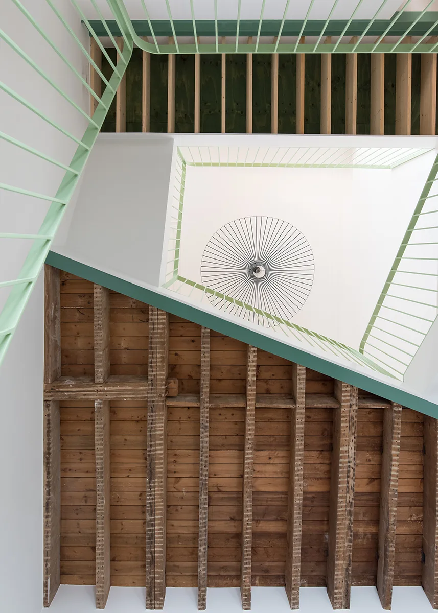 A low-angle view looking up a stairwell, showing exposed wooden ceiling joists, pale green metal railings, and a circular spoked pendant light.
