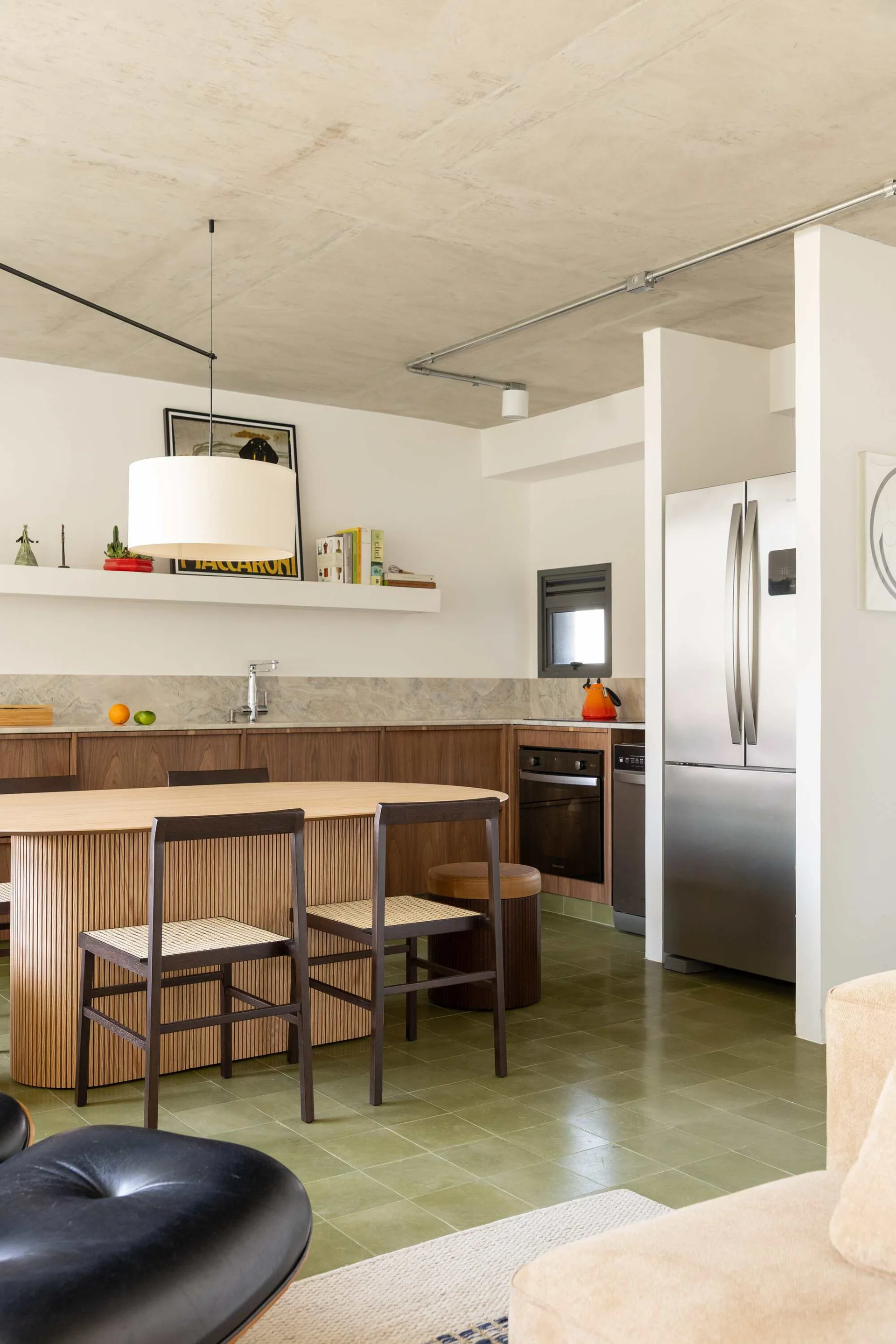 A modern kitchen with olive green tiled floors, wood cabinetry, a concrete ceiling, and an oval dining table with woven chairs.