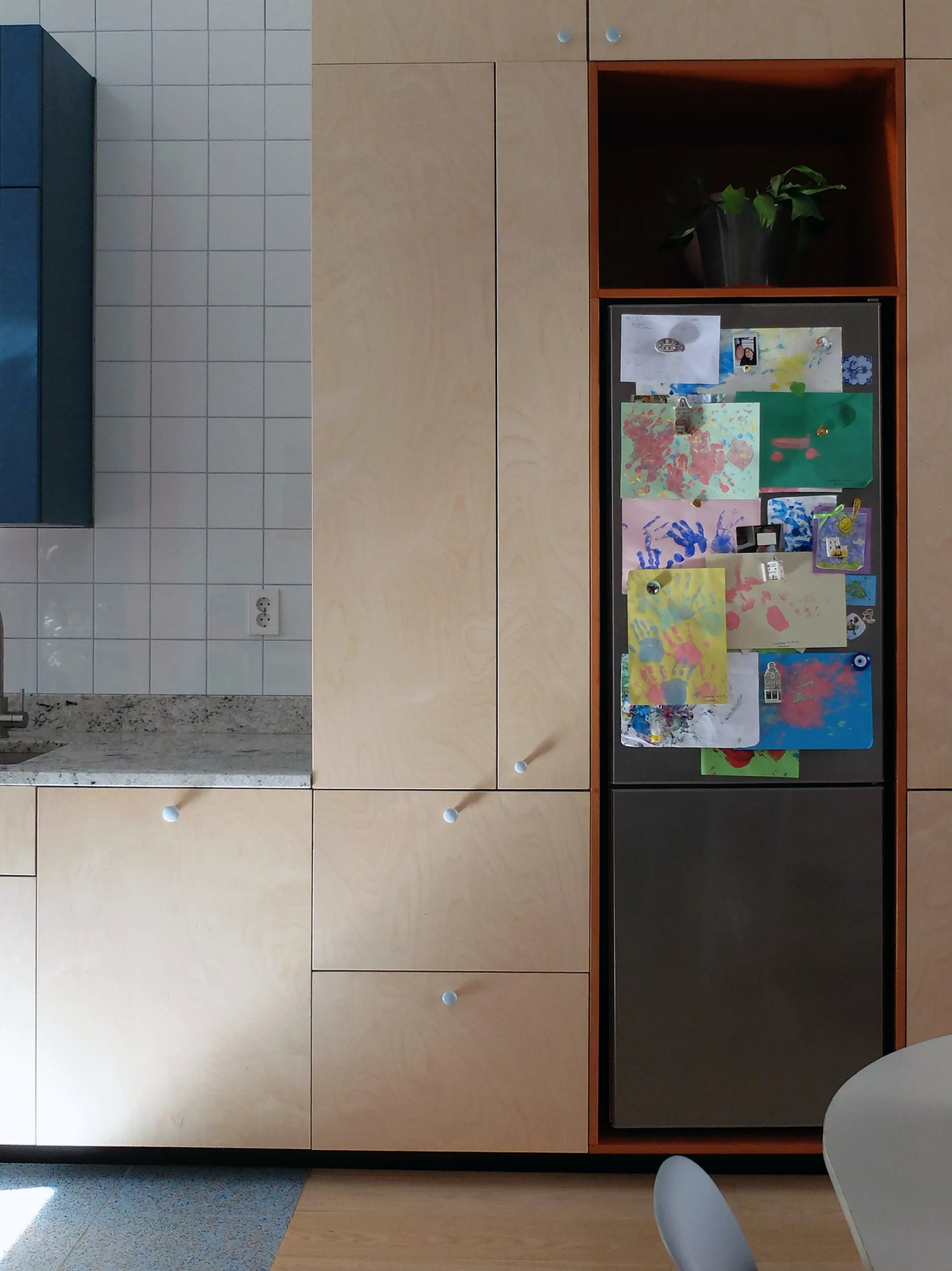 Modern kitchen with built-in light plywood cabinets enclosing a refrigerator decorated with children's artwork next to white grid backsplash tiles.