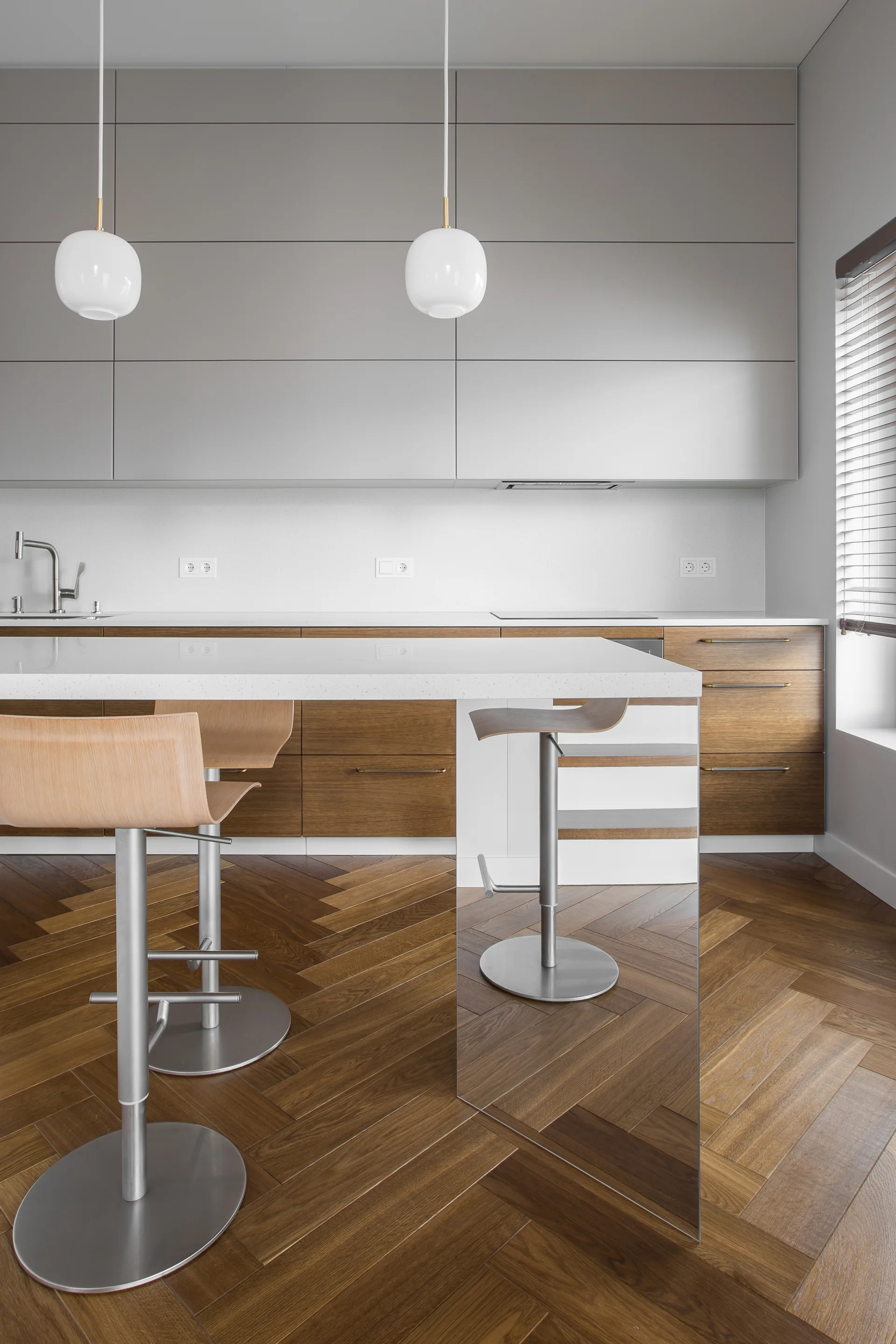 A modern kitchen featuring grey and wood cabinets, a white countertop island with wooden bar stools, and herringbone wood flooring.
