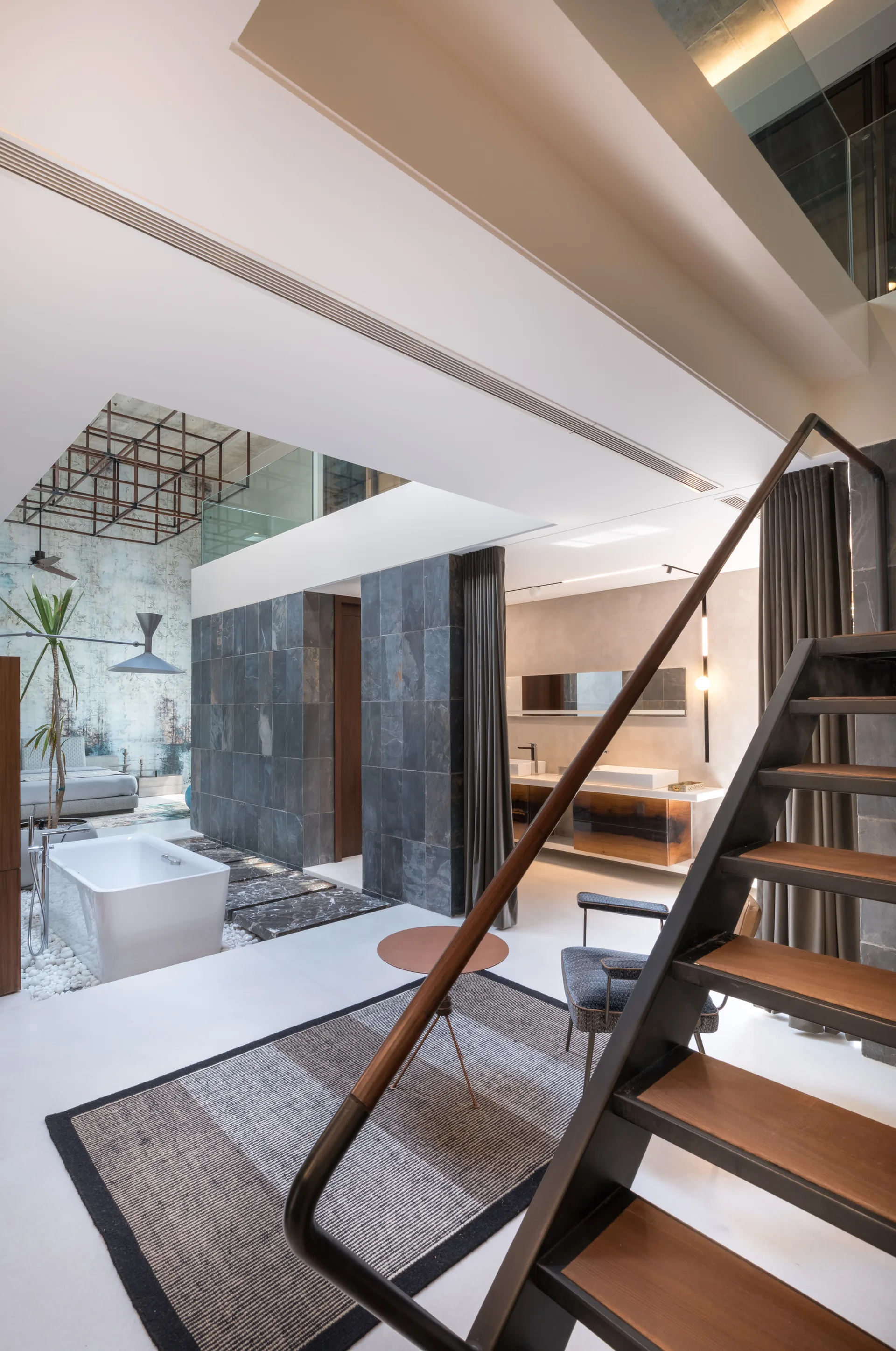 An open-concept bathroom features a freestanding tub on white pebbles, dark stone partition walls, and an open-tread staircase in the foreground.