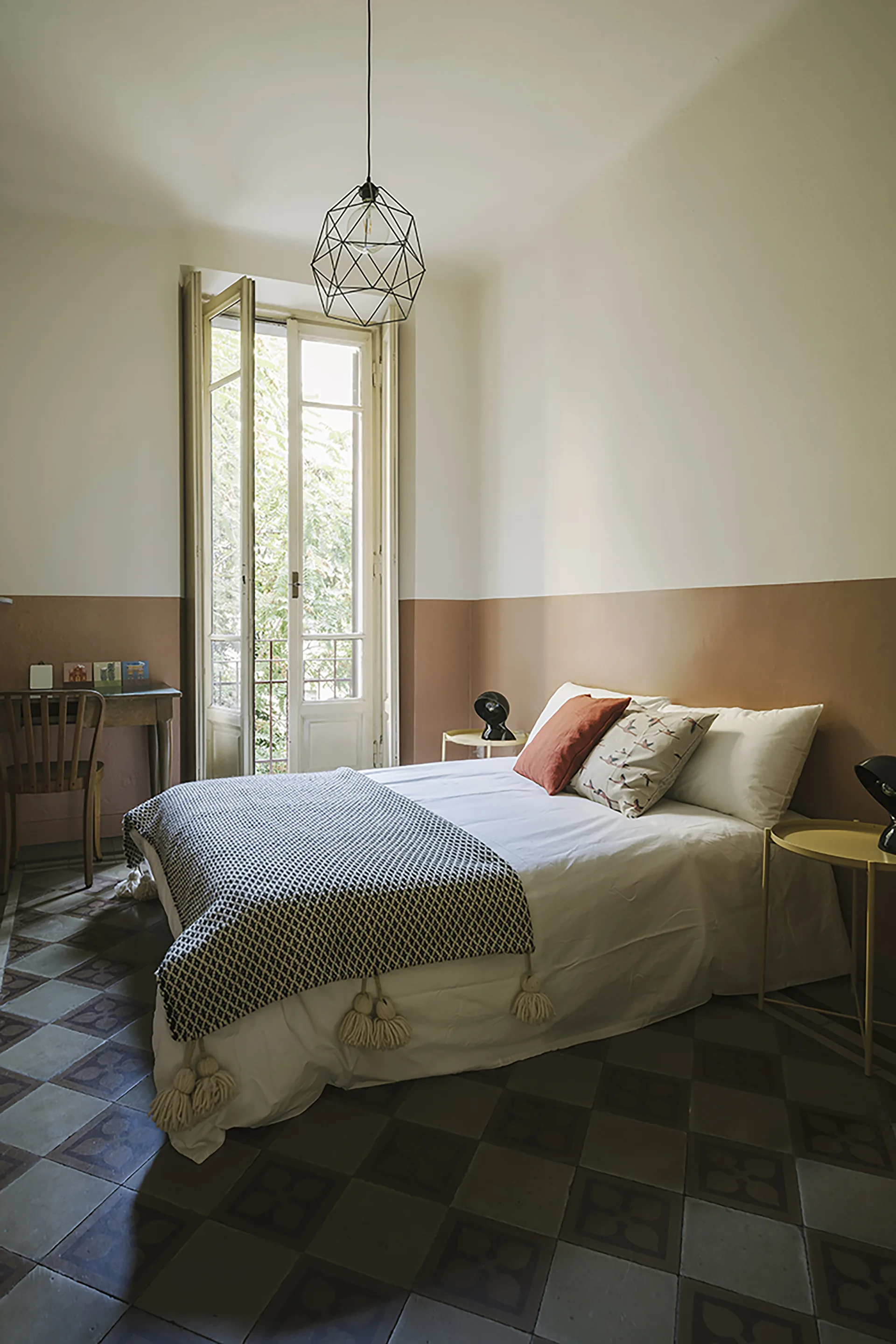 A bedroom featuring a bed with white linens and a tasseled throw blanket, set against walls painted white and terracotta, over patterned tile floors.