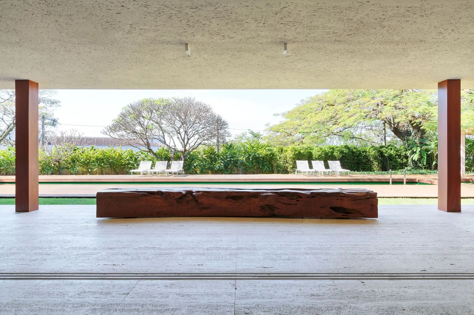 A minimalist covered patio with stone-tiled flooring, wood support columns, and a large rustic log bench facing an outdoor pool.