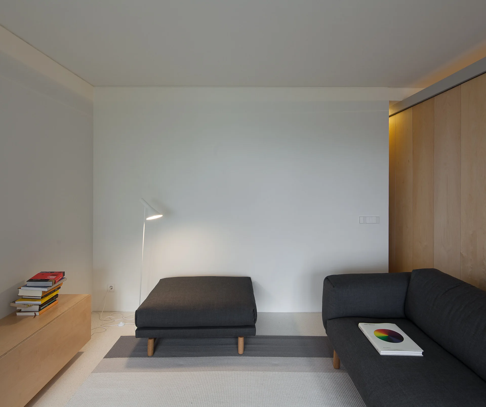 A minimalist living room features a dark gray sofa, matching ottoman, a slender white floor lamp, and a light wood paneled wall.