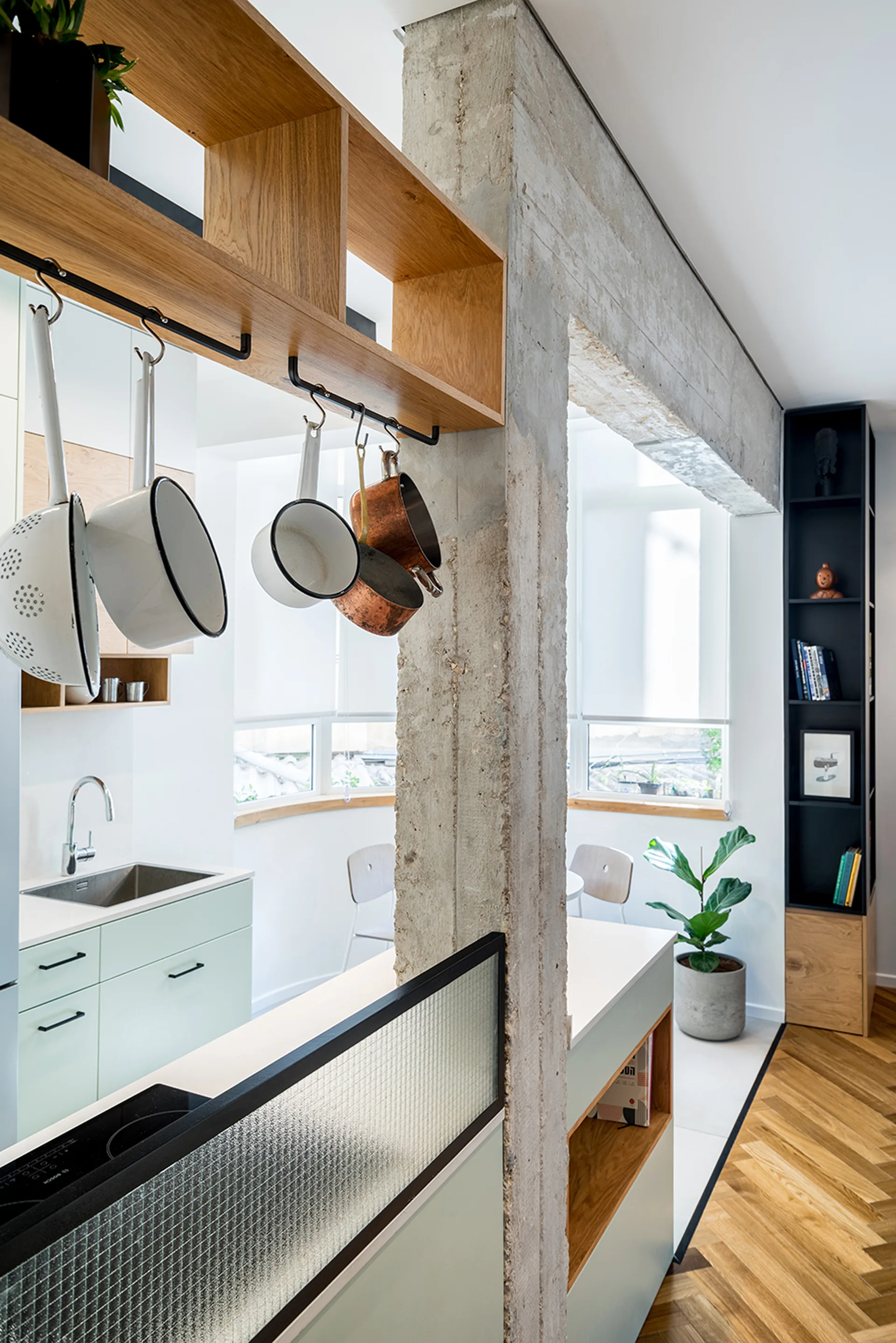 A modern kitchen featuring pale green cabinets, a raw concrete structural column, and enamel pots hanging from a wooden shelf.