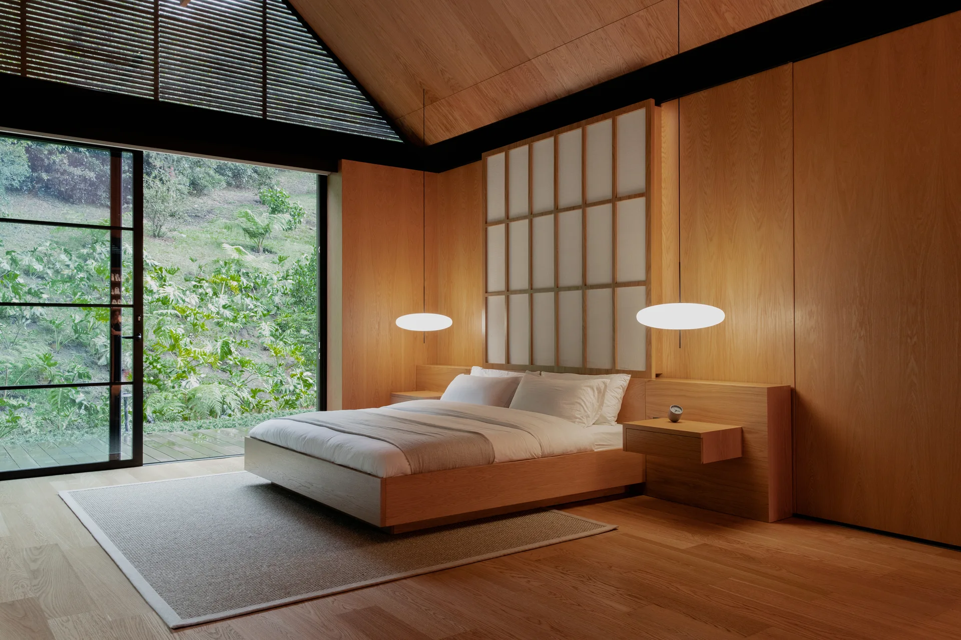 A minimalist bedroom featuring a low-profile bed with a shoji-style headboard, wood-paneled walls, and sliding glass doors that open to a garden.
