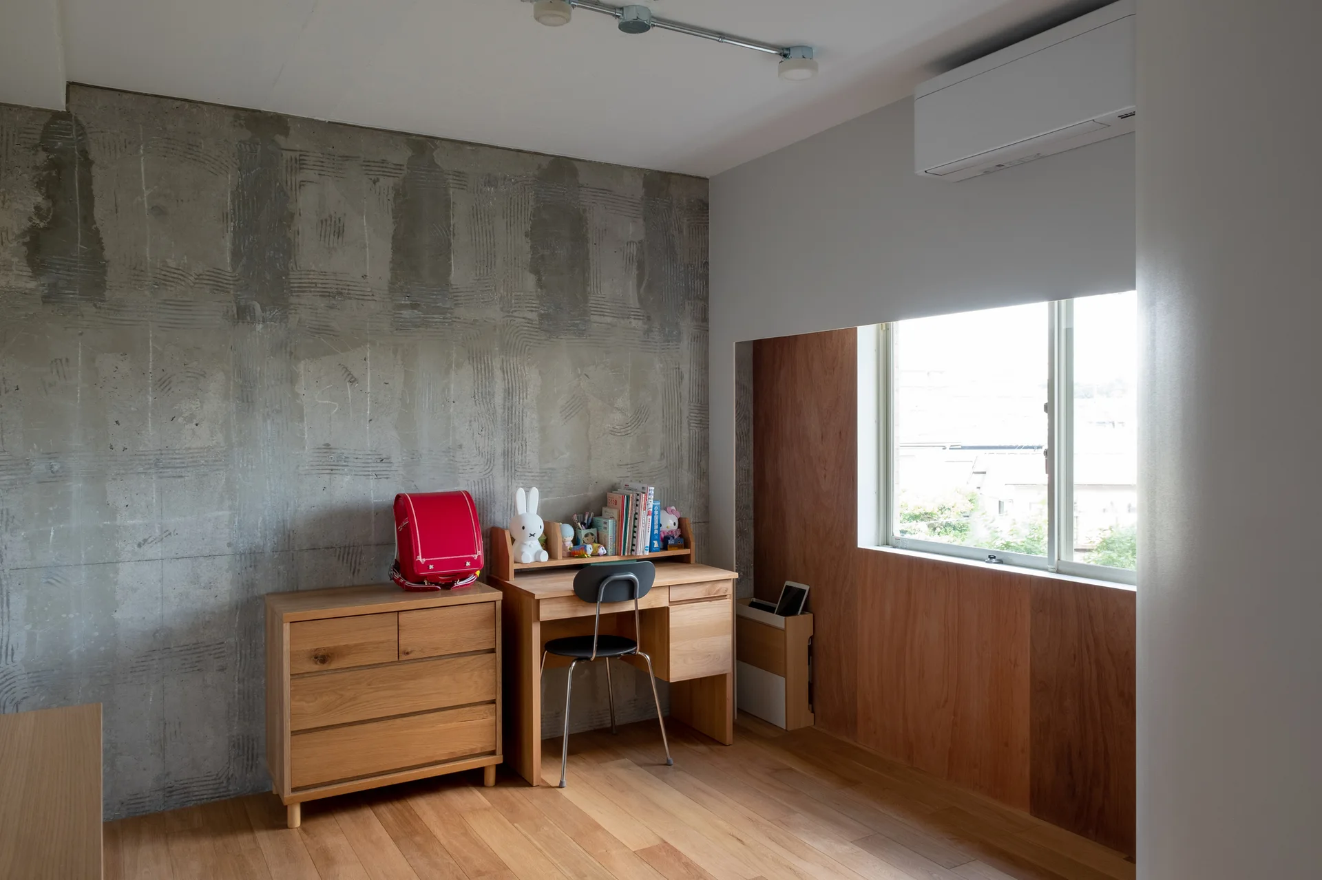 A minimalist kids study corner featuring a wooden desk and drawers against a textured concrete wall and a window surrounded by wood panels.