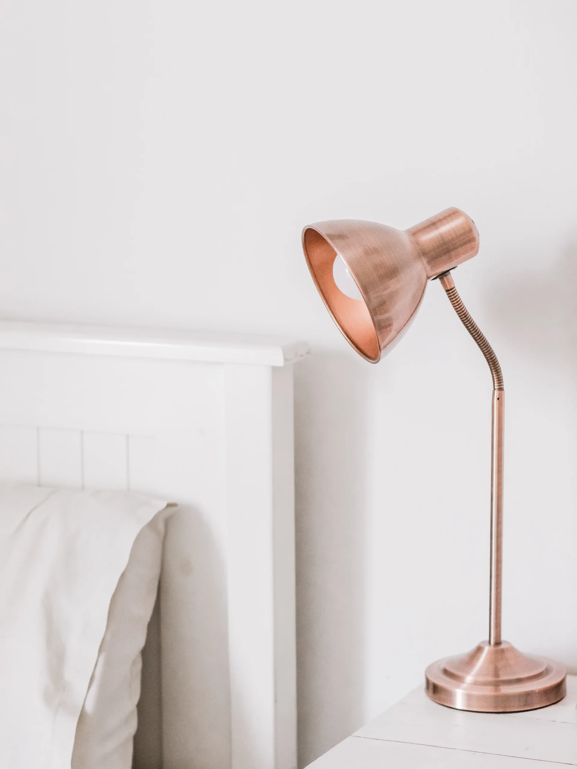 A brushed copper gooseneck lamp sits on a white bedside table next to a matching white wooden headboard against a plain white wall.