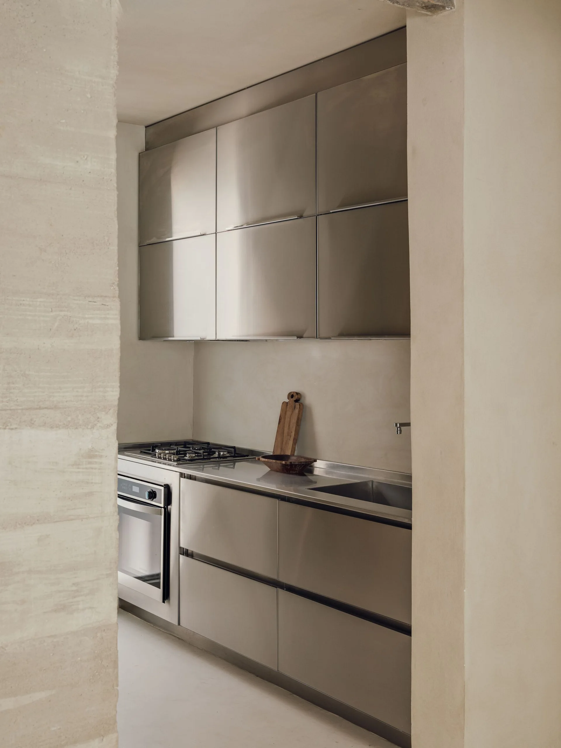 A recessed kitchen features brushed stainless steel cabinets, integrated appliances, and a wood cutting board set against smooth beige plaster walls.