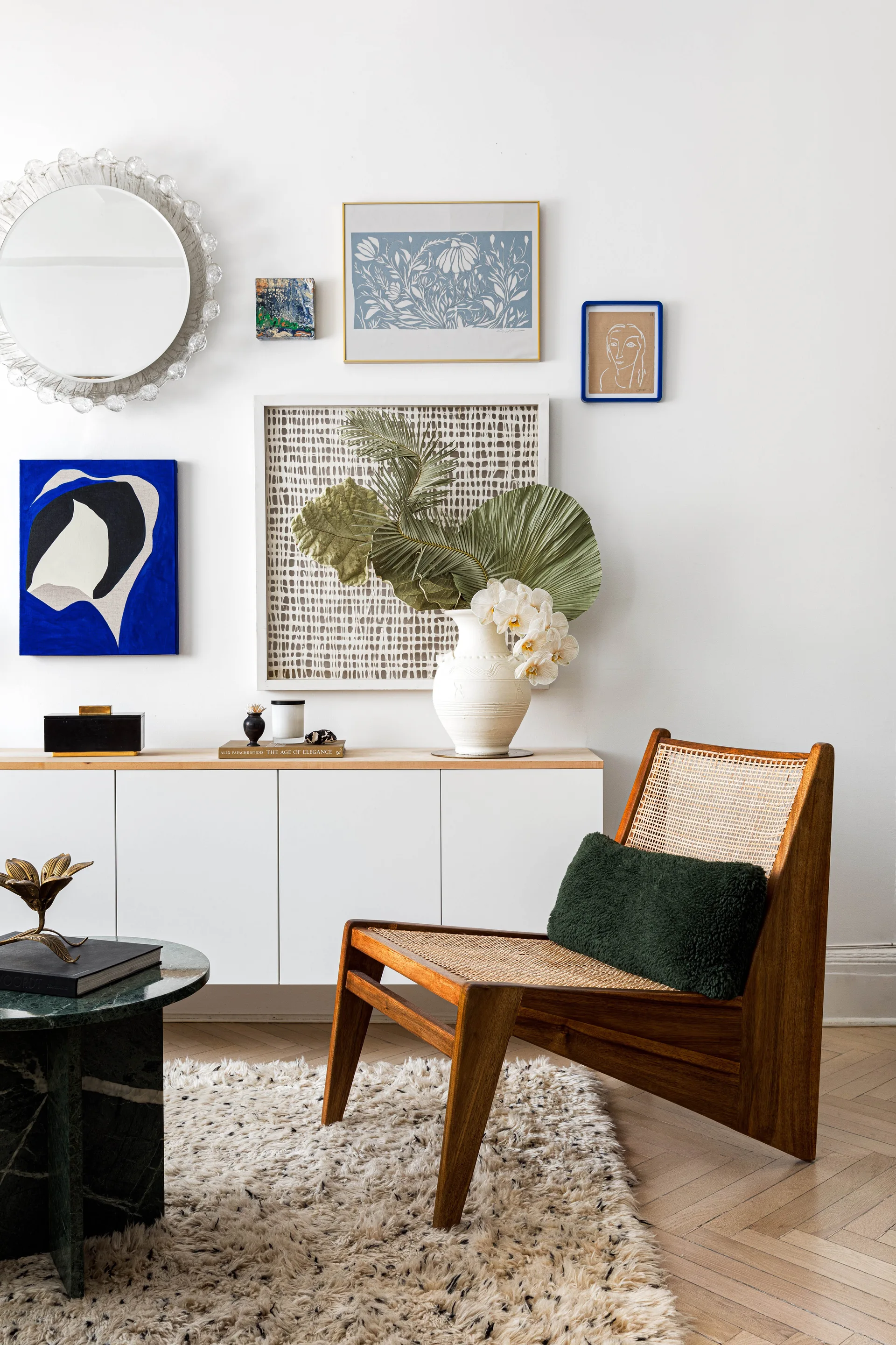 A mid-century wood and cane lounge chair sits near a white console table with a gallery wall of art and a round mirror.