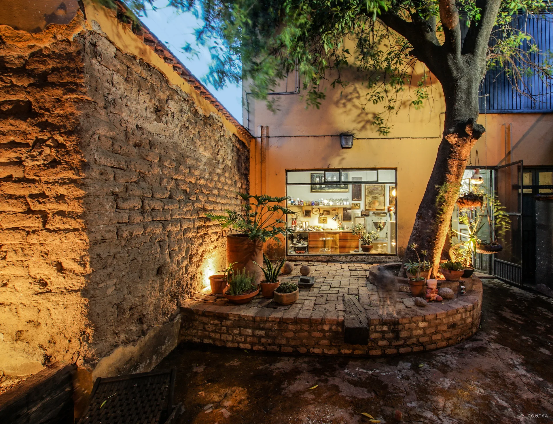 A rustic brick courtyard featuring a curved raised brick planter with a large tree, terracotta potted plants, and a window viewing a lit studio.