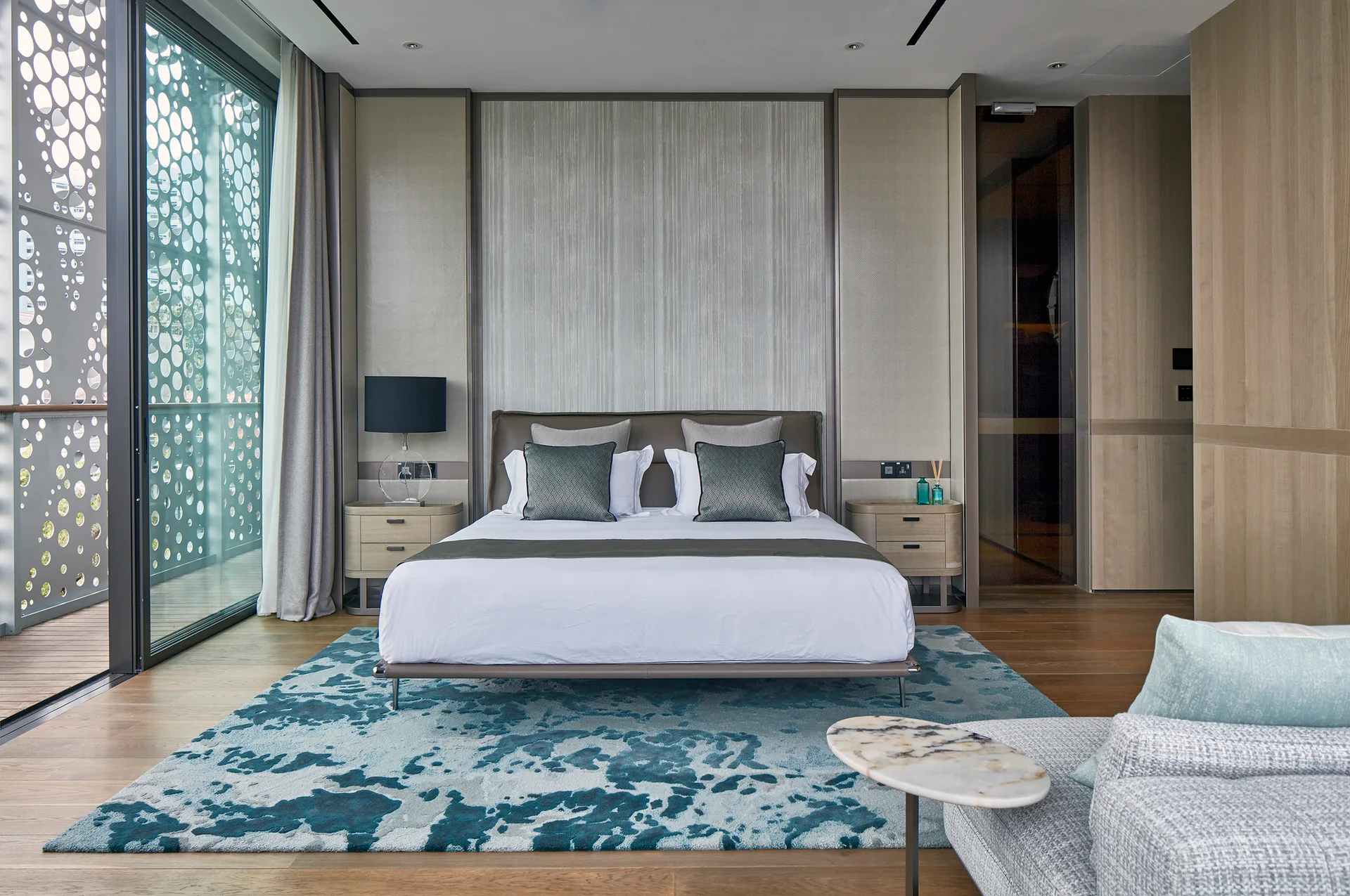 A modern bedroom features a king-sized bed, textured beige wall panels, a blue abstract area rug, and a large sliding glass door with a decorative screen.