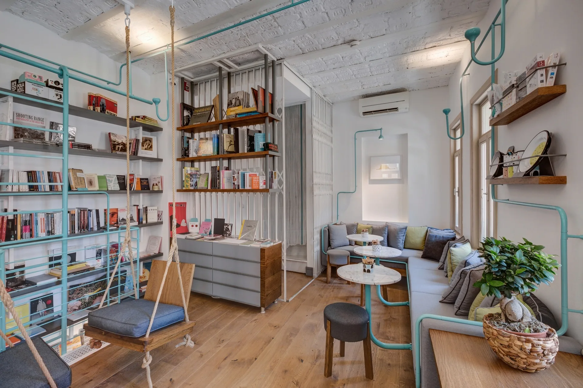A bright interior lounge featuring a grey sectional sofa, white brick ceiling, bookshelves supported by teal pipes, and a suspended wooden swing.
