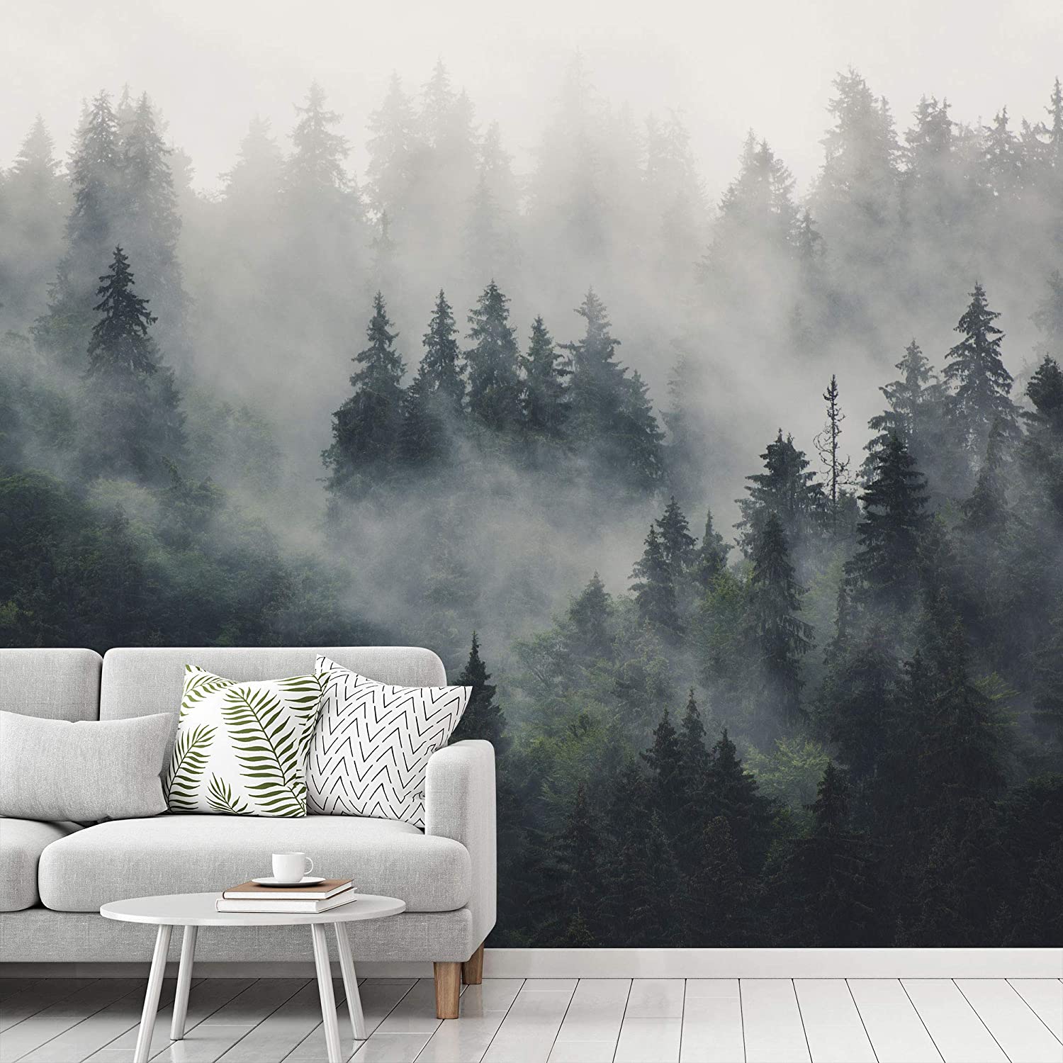 A light gray sofa with patterned throw pillows sits in front of a large wall mural depicting a foggy pine forest over white wood plank floors.