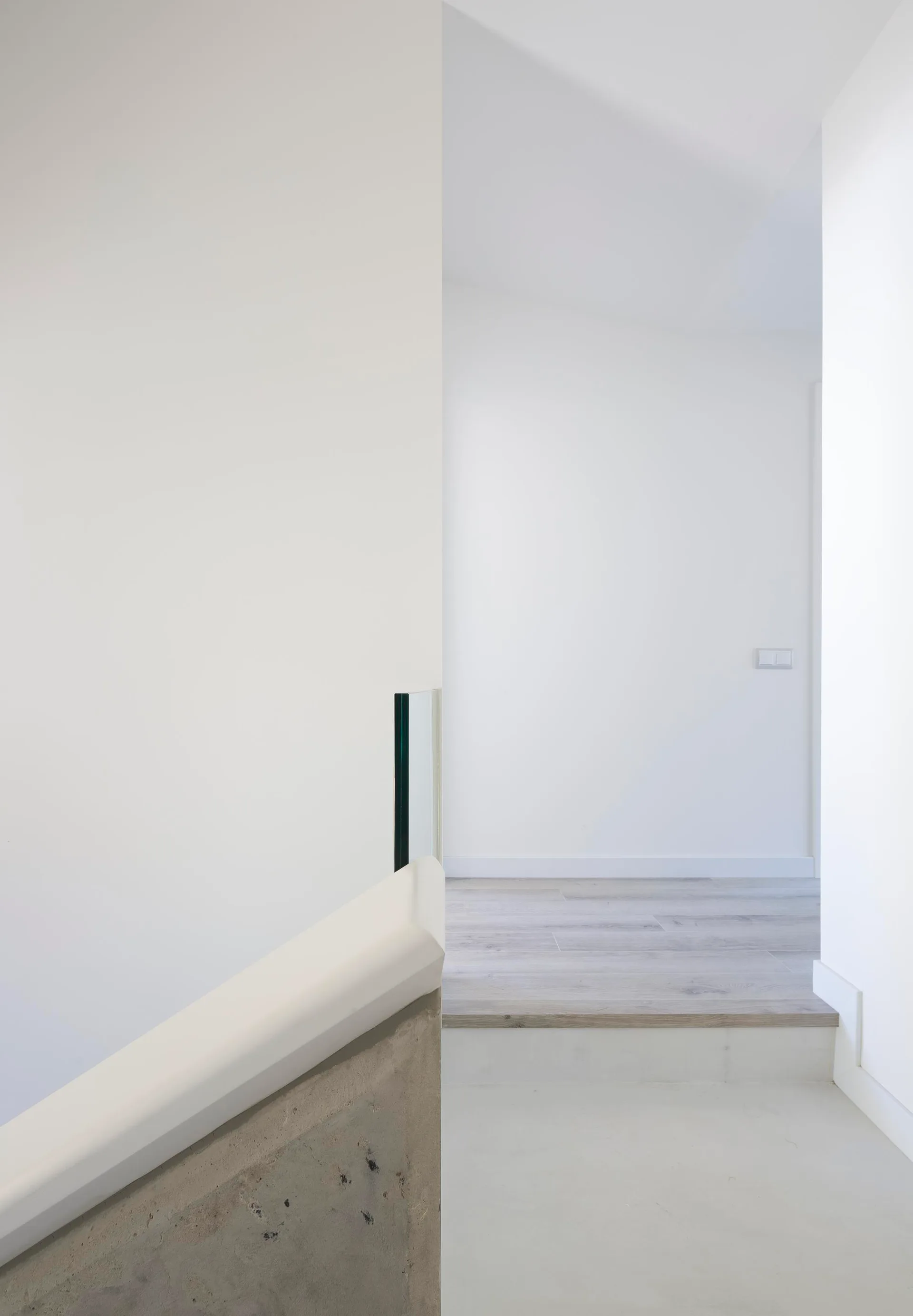 A modern minimalist staircase landing with smooth white walls, a simple handrail, and light wood plank flooring.