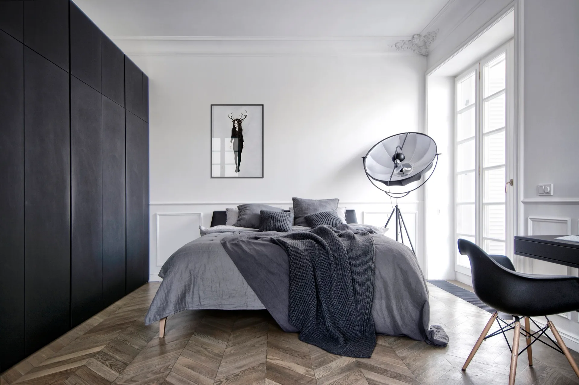 A modern minimalist bedroom featuring a gray bed, a black built-in wardrobe, herringbone wood floors, and a studio tripod lamp.