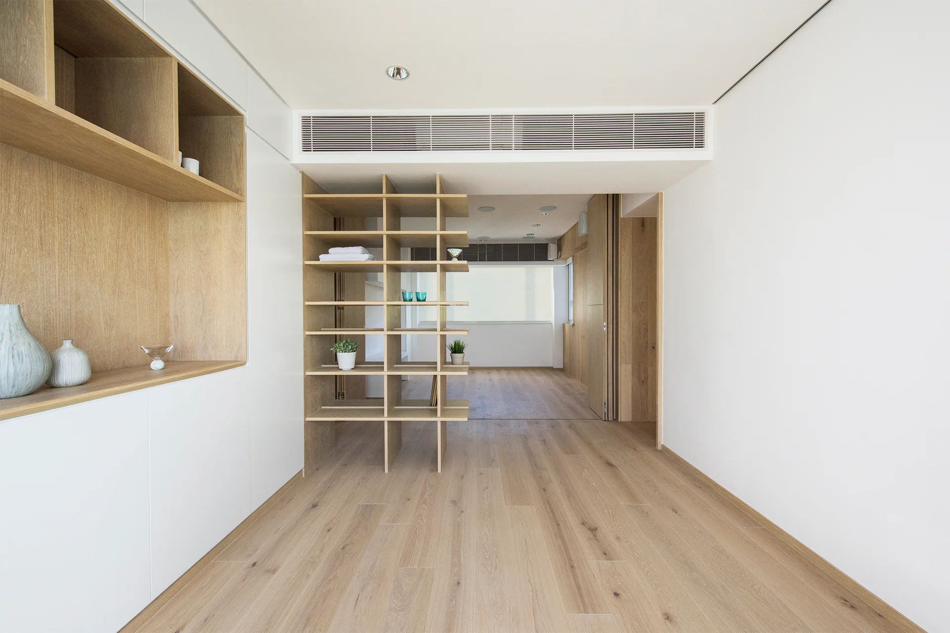 An airy minimalist room features light wood plank flooring, white built-in cabinets, and an open wood grid shelving unit serving as a partition.