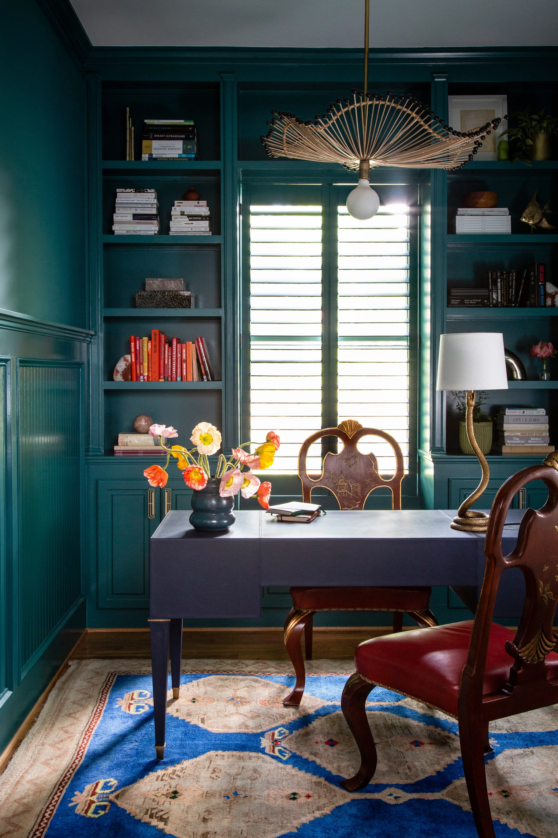 A home office featuring deep teal painted built-in bookshelves, a navy desk with a floral arrangement, and a patterned blue rug.