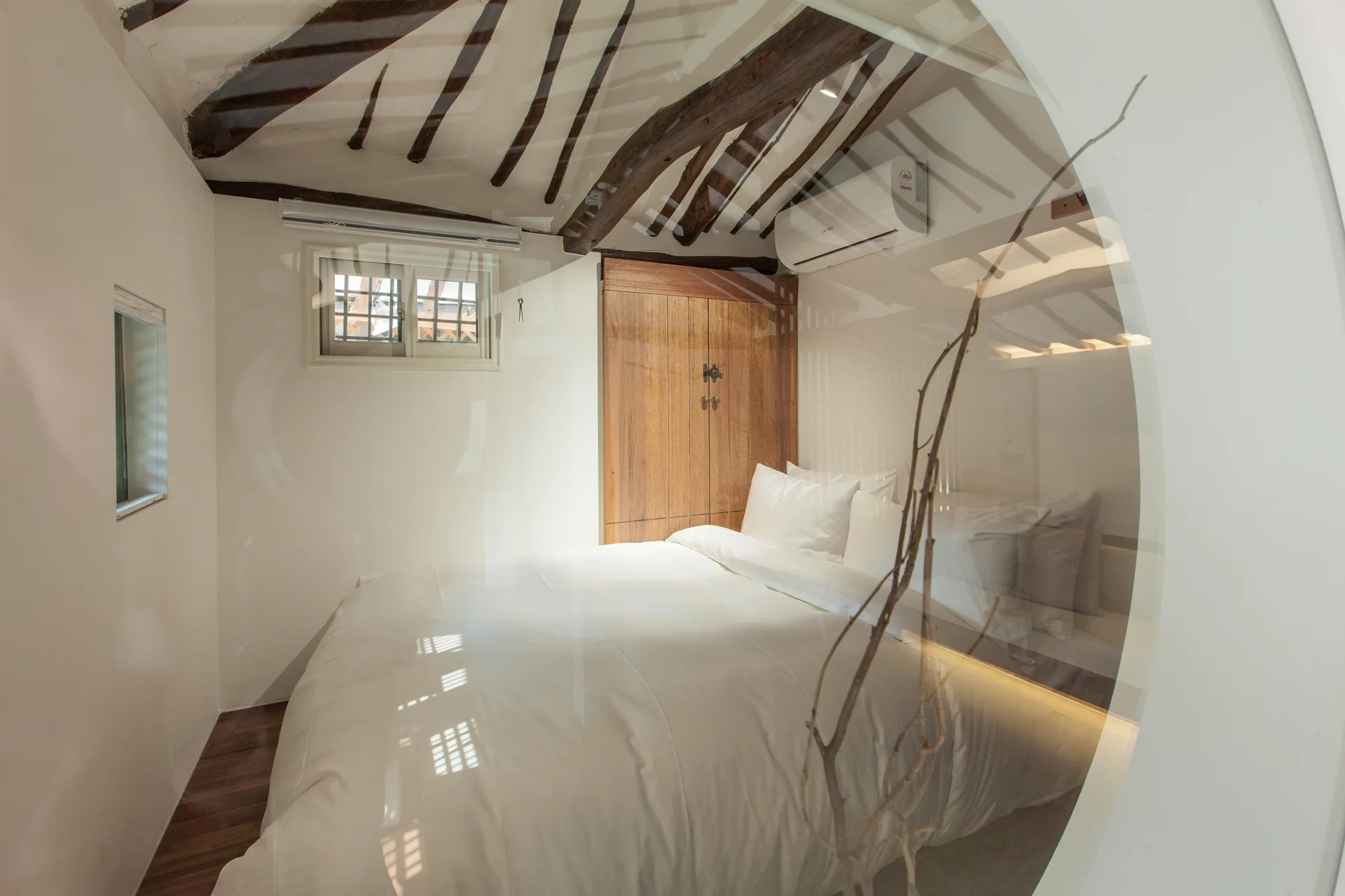 A minimalist bedroom featuring a white bed, a wooden wardrobe, and exposed dark wood ceiling beams, viewed through a circular glass partition.