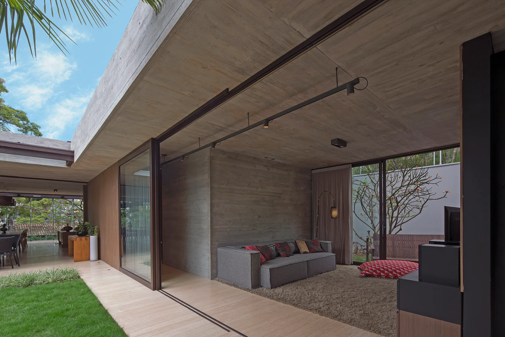 A modernist living room features raw concrete walls, a gray sectional sofa, a shag rug, and large sliding glass doors leading to a green lawn.