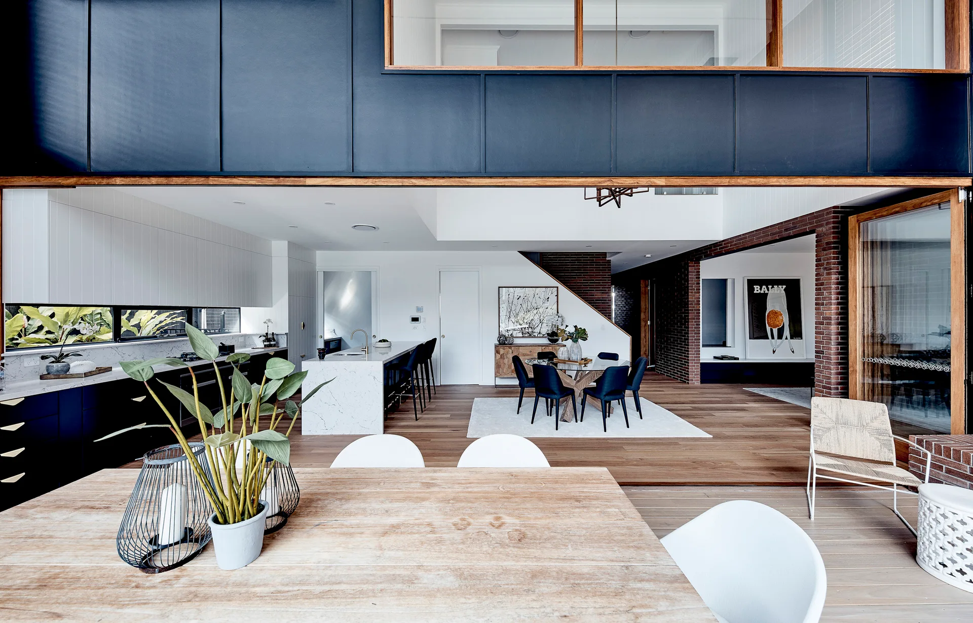 An open-concept kitchen and dining area features black cabinetry, a white marble kitchen island, exposed brick walls, and light wood flooring.