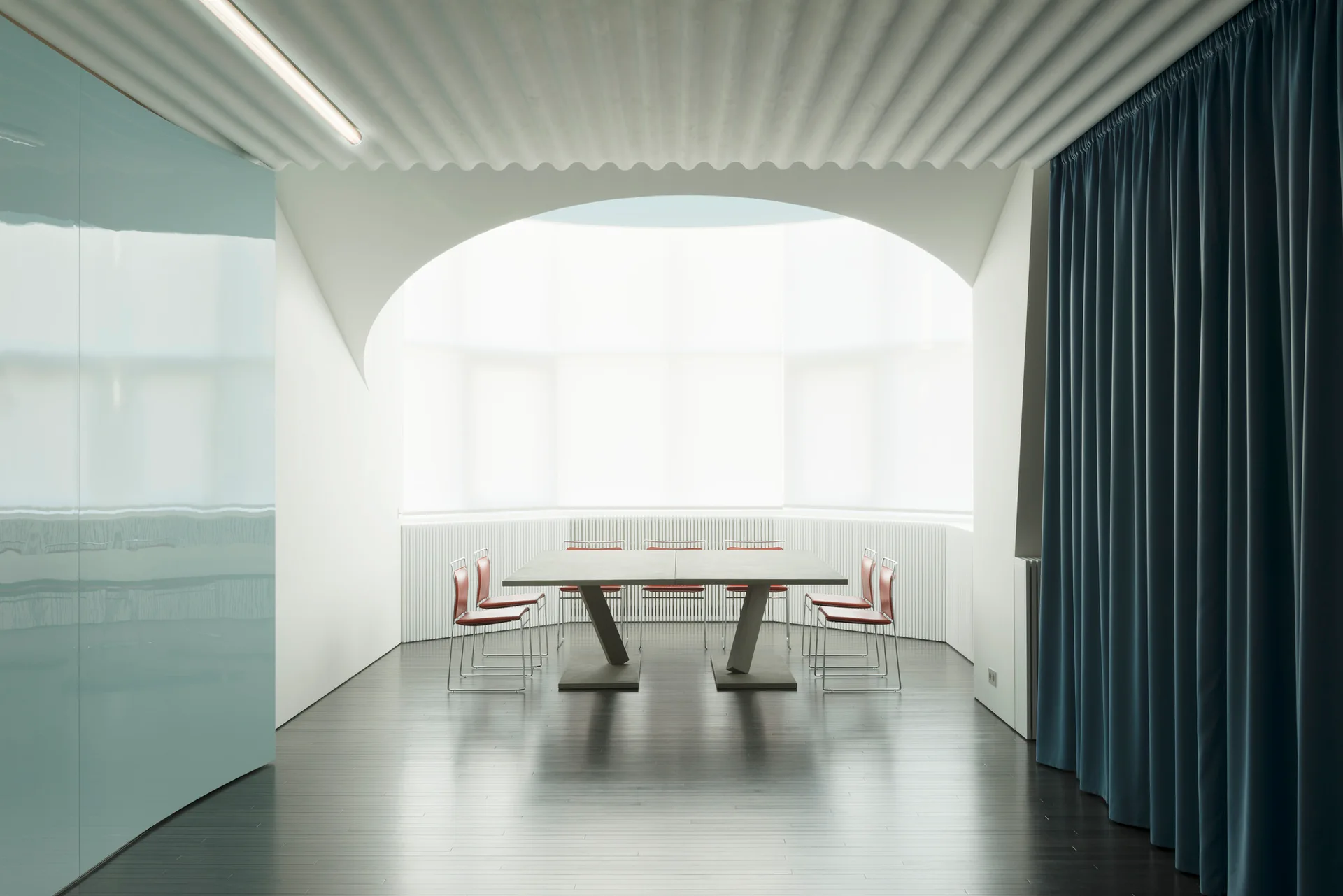 A minimalist dining room features a central rectangular table with red chairs beneath a large white archway, beside a heavy blue curtain.