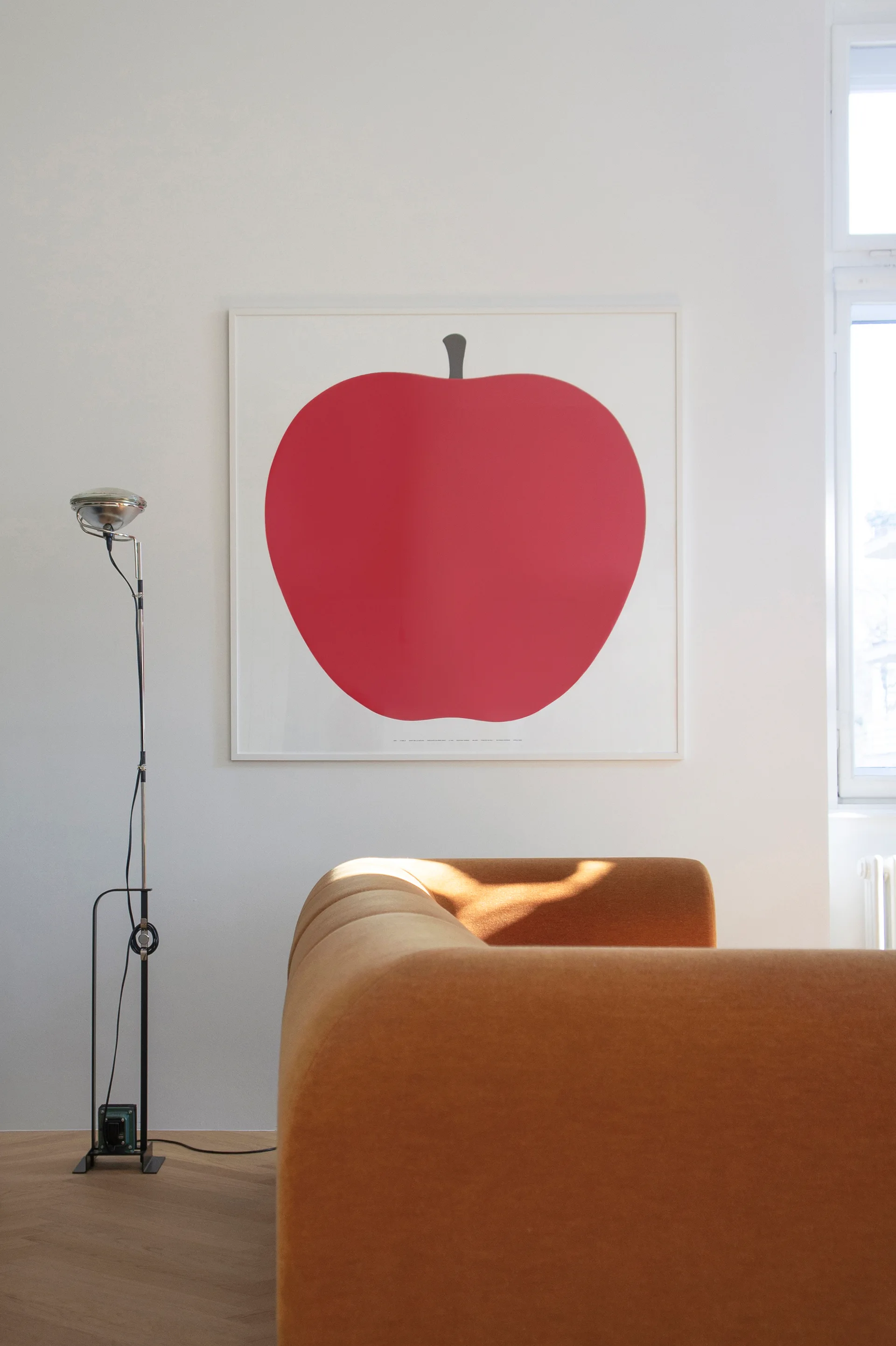 A modern tan sofa sits in front of a white wall decorated with a large framed print of a red apple.