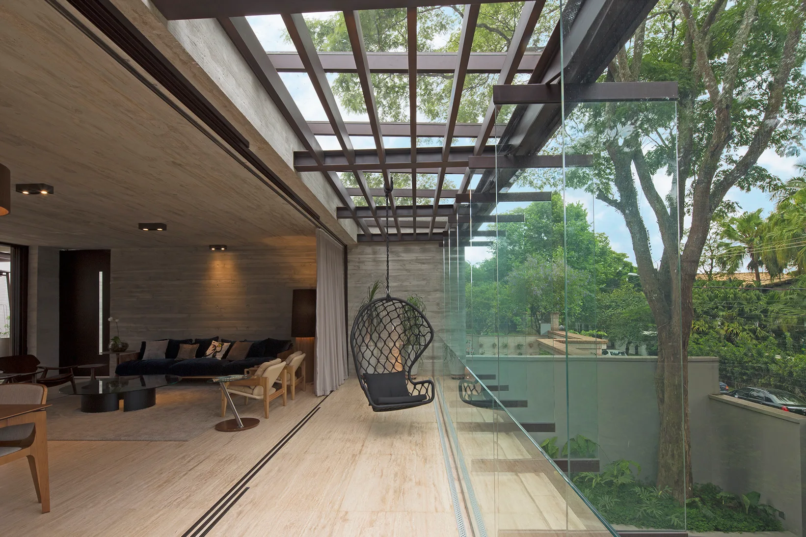 A modern indoor-outdoor terrace with stone tile flooring, a suspended black lounge chair, a metal pergola, and a frameless glass railing.