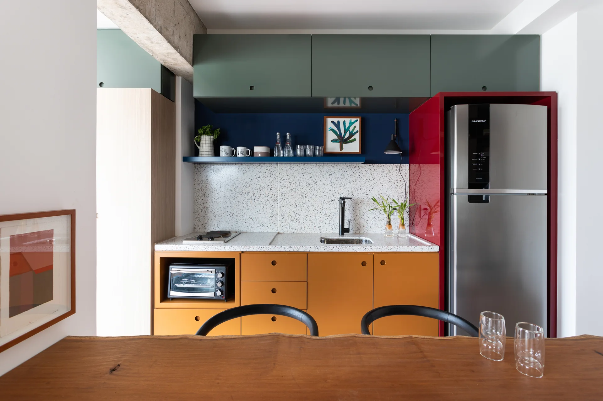 A modern kitchen features green and mustard color-blocked cabinets, a speckled terrazzo backsplash, and a stainless steel refrigerator framed in red.