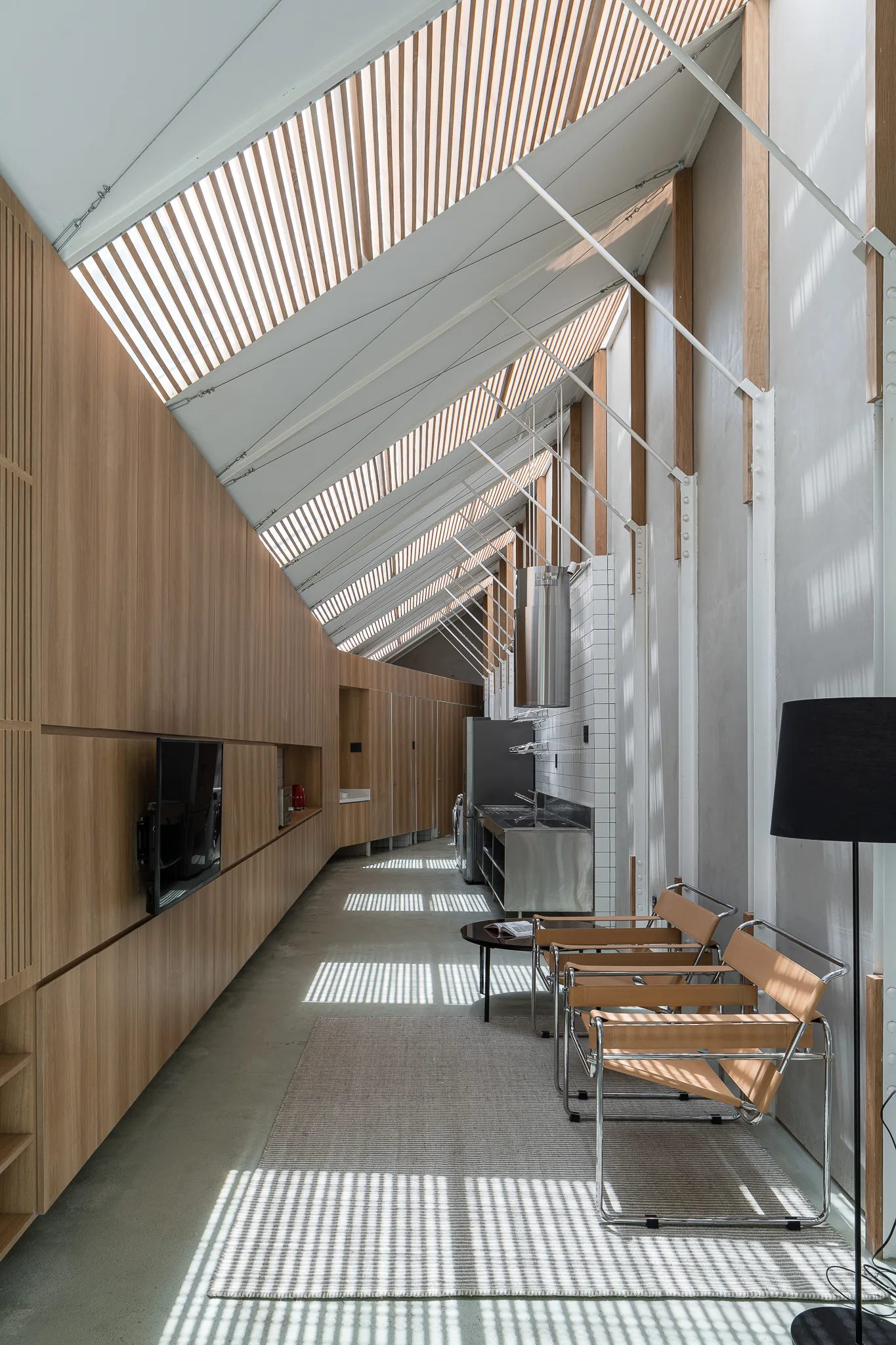 A modern elongated kitchen and seating area features a sloped wood louvered ceiling, a concrete floor, leather chairs, and wood paneling.