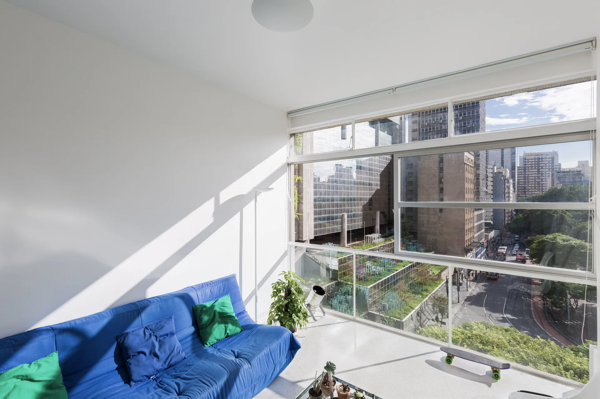 A minimalist living room features a bright blue sofa against a white wall next to a large window overlooking a city street.