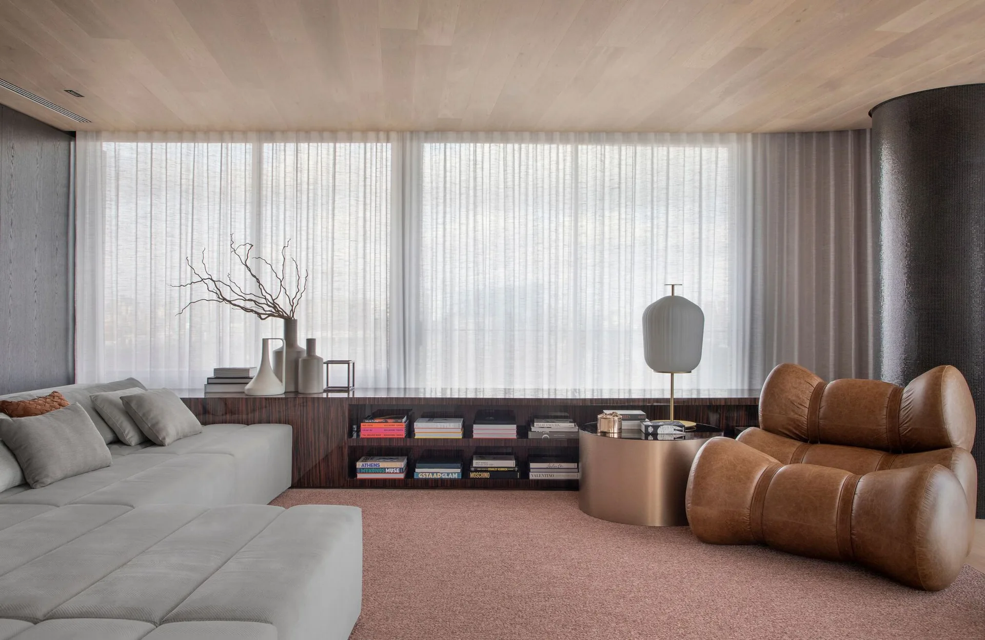 A contemporary living room with a grey sectional, a bulbous brown leather armchair, and a low wooden console in front of sheer curtains.