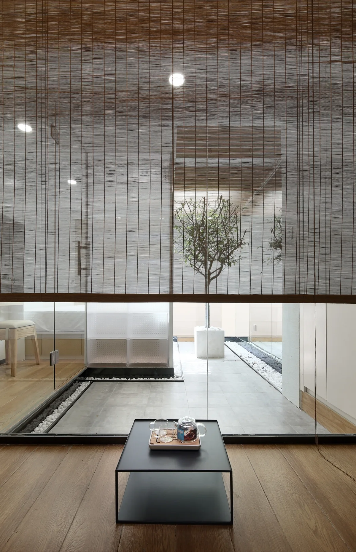 A minimalist seating area with wood floors and a low black tea table looks out onto a glass-enclosed indoor courtyard containing a small tree.