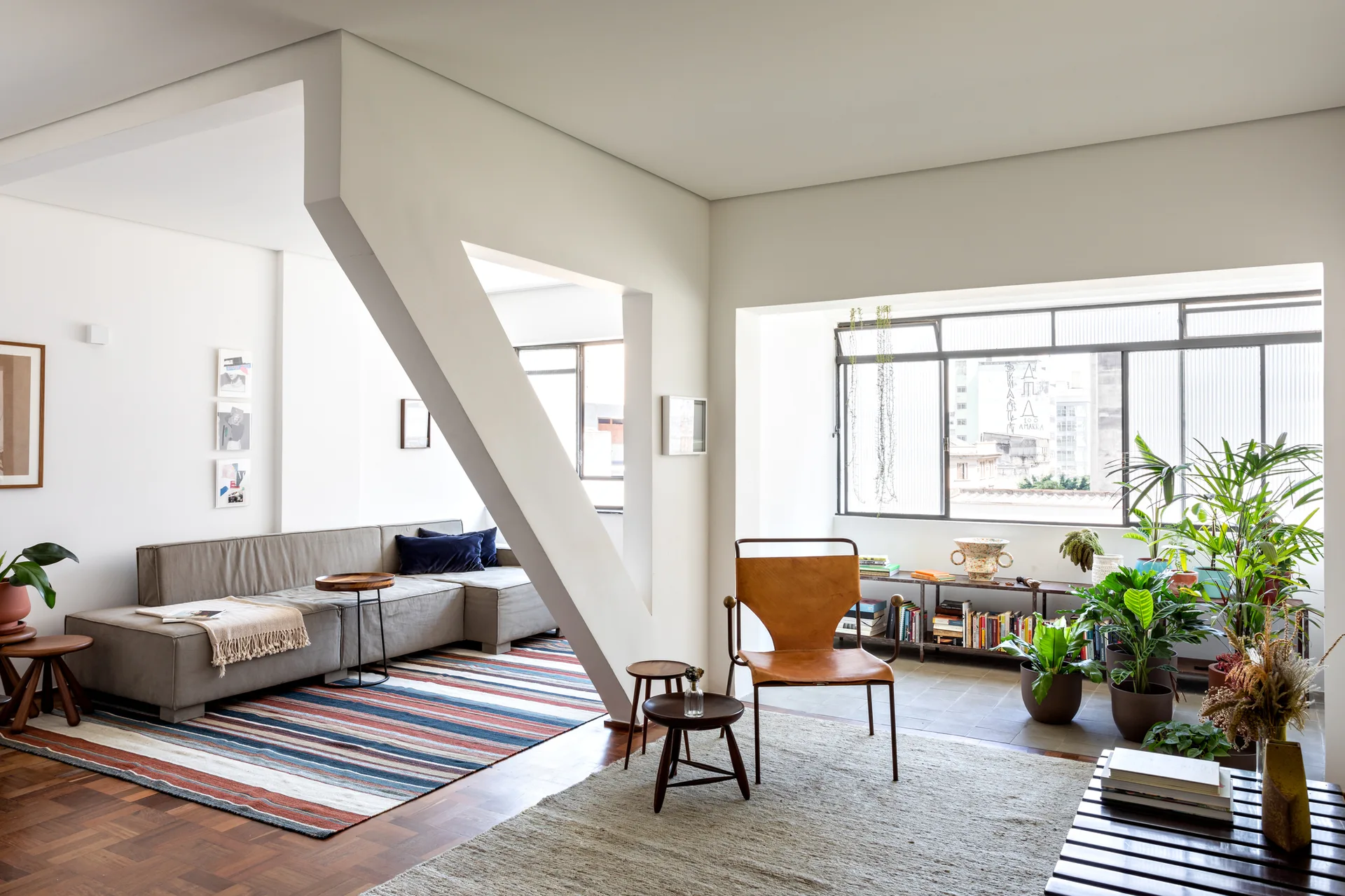 A light-filled living room featuring an angled white support beam, a grey sofa on a striped rug, a leather sling chair, and potted plants.