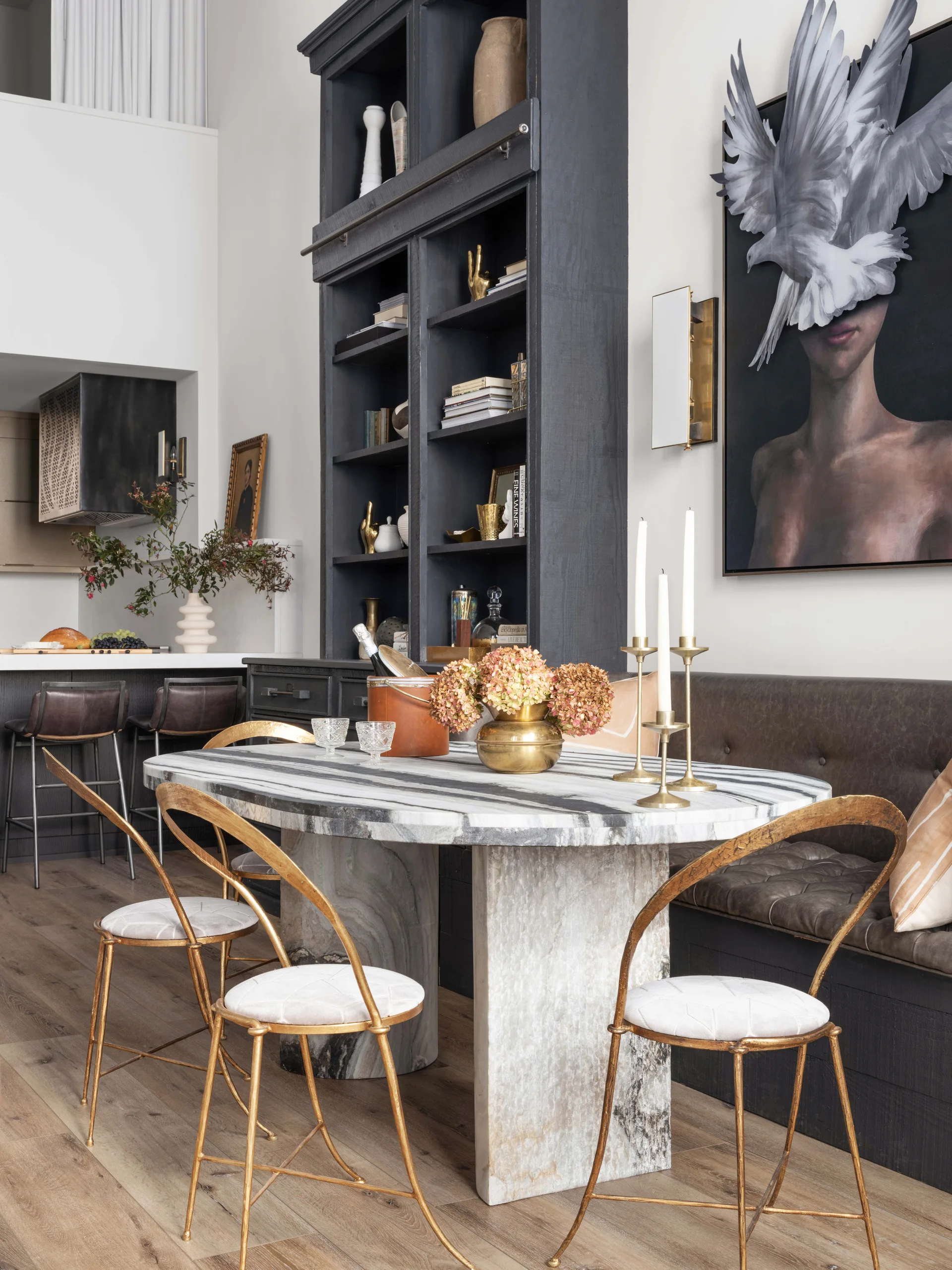 An oval veined marble dining table is surrounded by three gold metal chairs and a dark tufted banquette in front of a tall black bookcase.