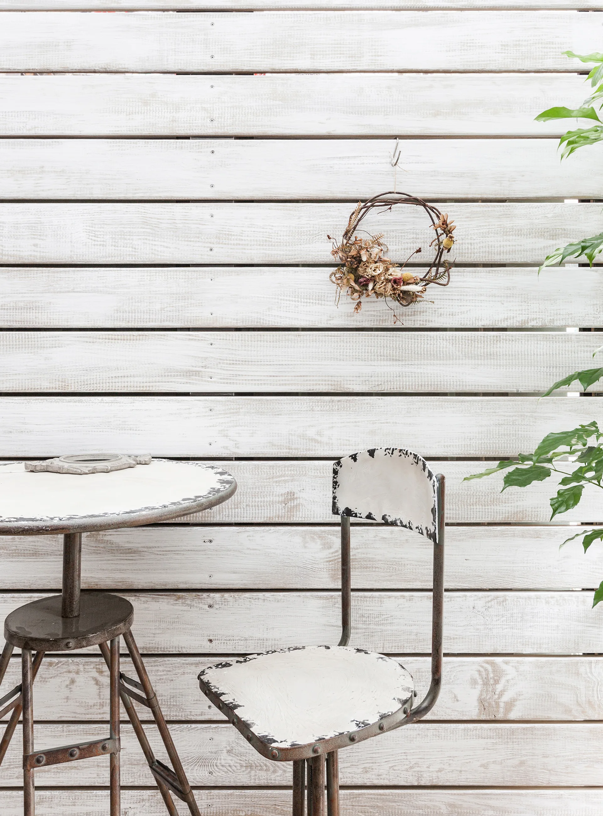 A distressed metal bistro table and chair stand before a white horizontal wood plank wall accented with a small dried twig wreath.